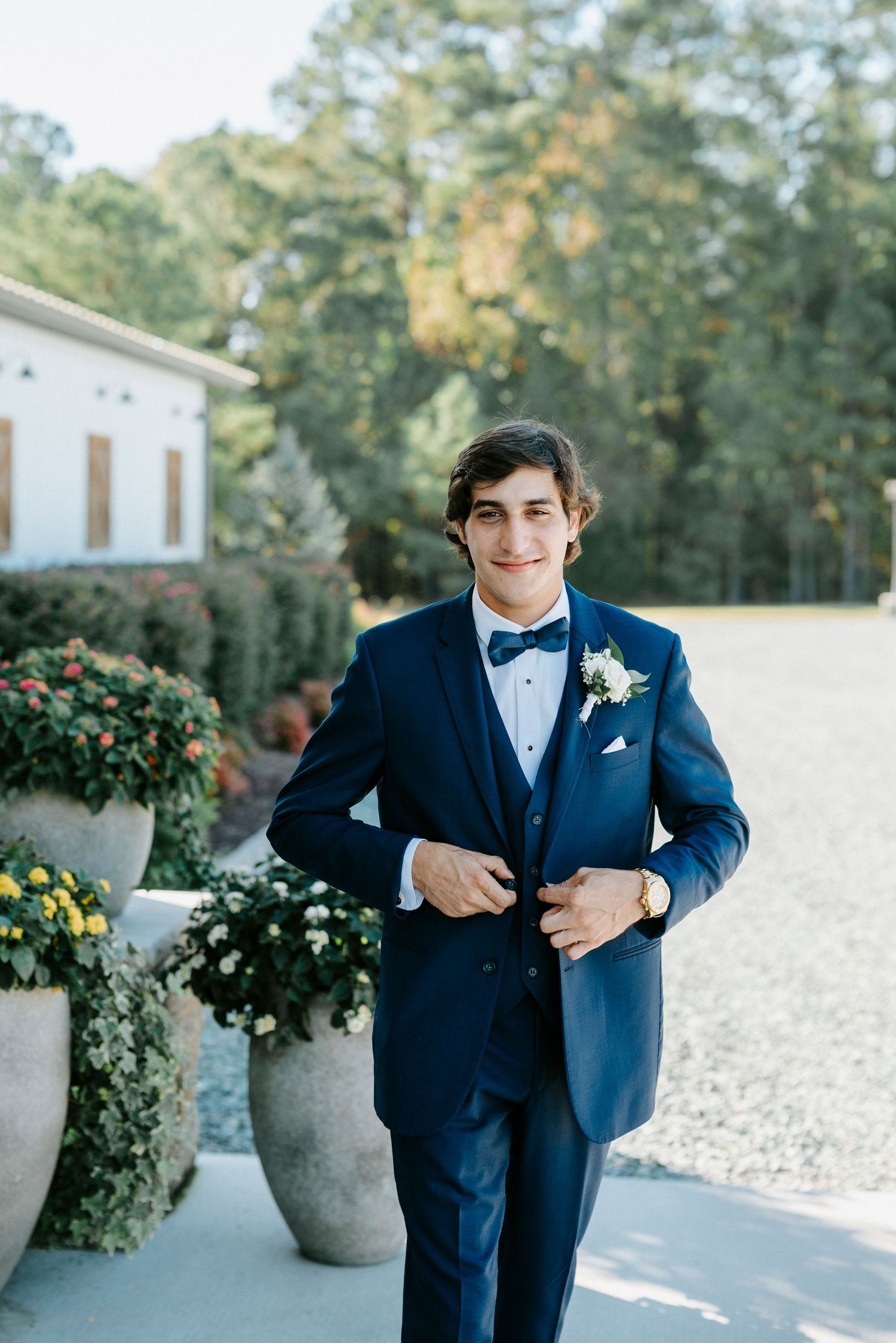 A groom in a navy blue tuxedo with a bowtie and boutonniere poses outdoors near decorative planters.