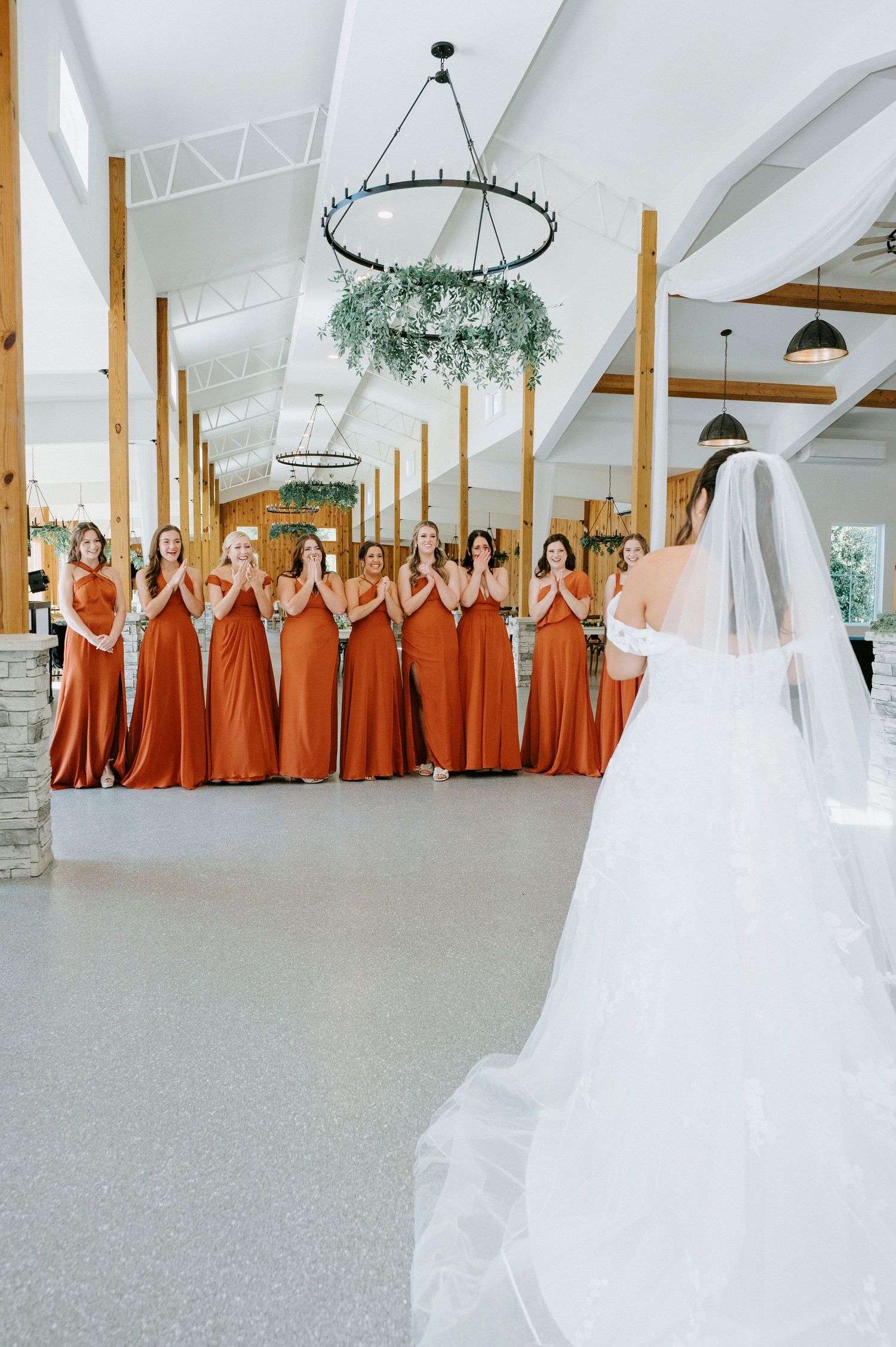 Bridesmaids in rust-colored dresses stand in a line while facing a bride in a white veil inside an elegant venue.