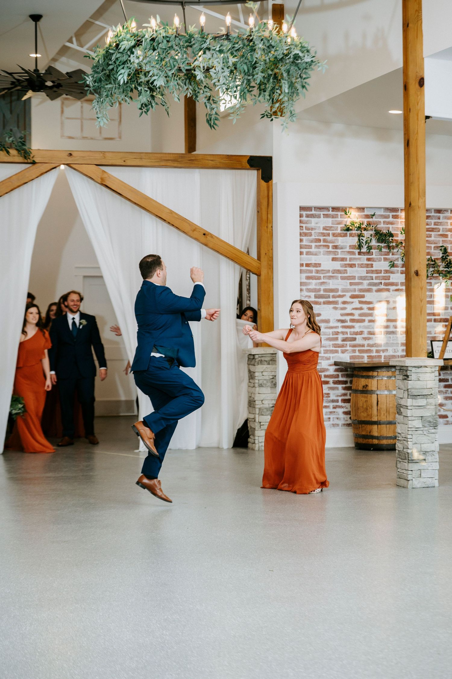 Wedding party members perform a fun synchronized jumping dance move in a bright white reception venue with brick accents.