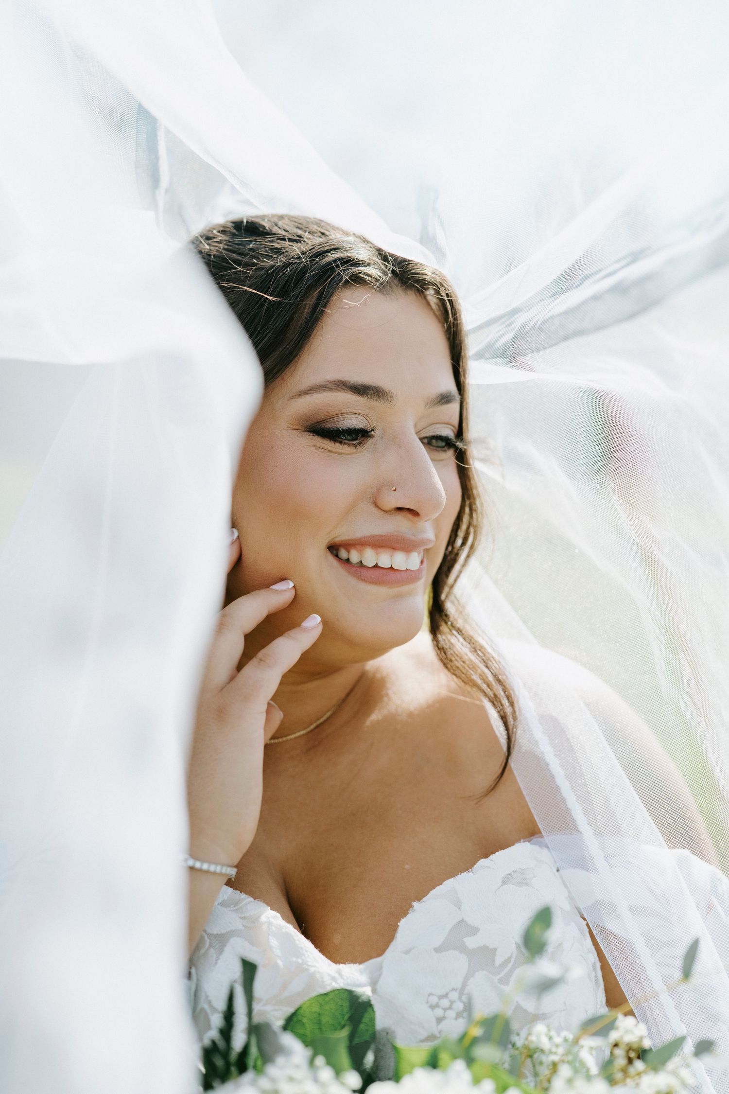 Bride in white veil smiles joyfully while holding delicate bouquet in soft ethereal lighting.