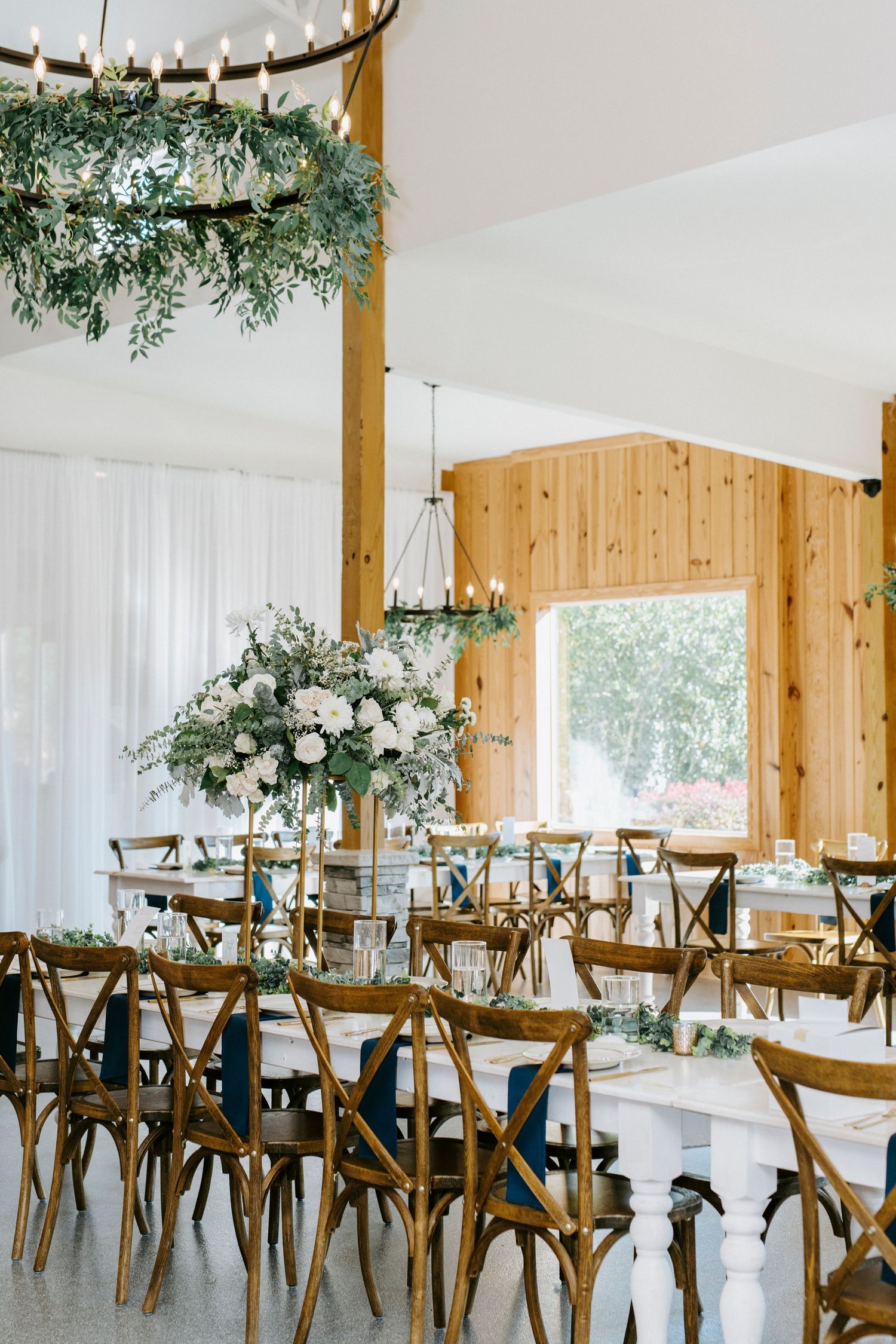 Rustic wedding reception venue with wooden cross-back chairs and greenery hanging from wooden beams.