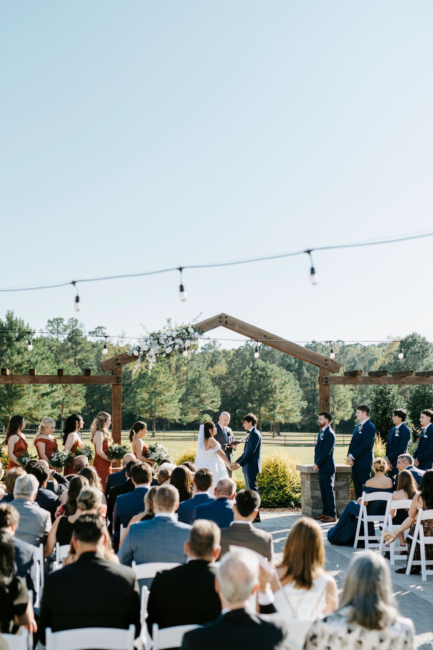 Outdoor wedding ceremony venue with guests seated under string lights and rustic wooden beams.