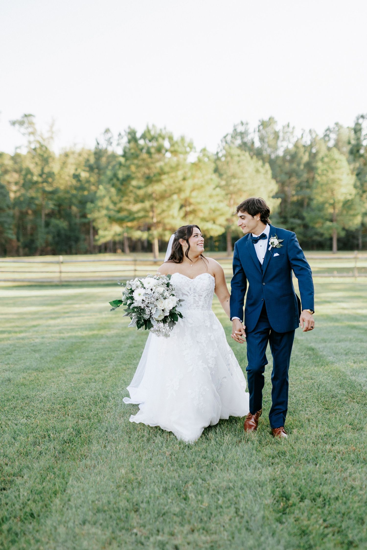 A couple in wedding attire walks hand in hand across a sunlit grassy field on their special day.
