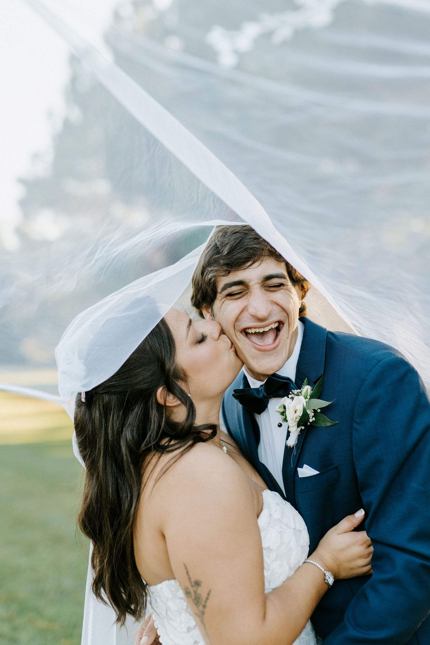 Joyful wedding couple laughing together outdoors with bride's veil flowing in the breeze.