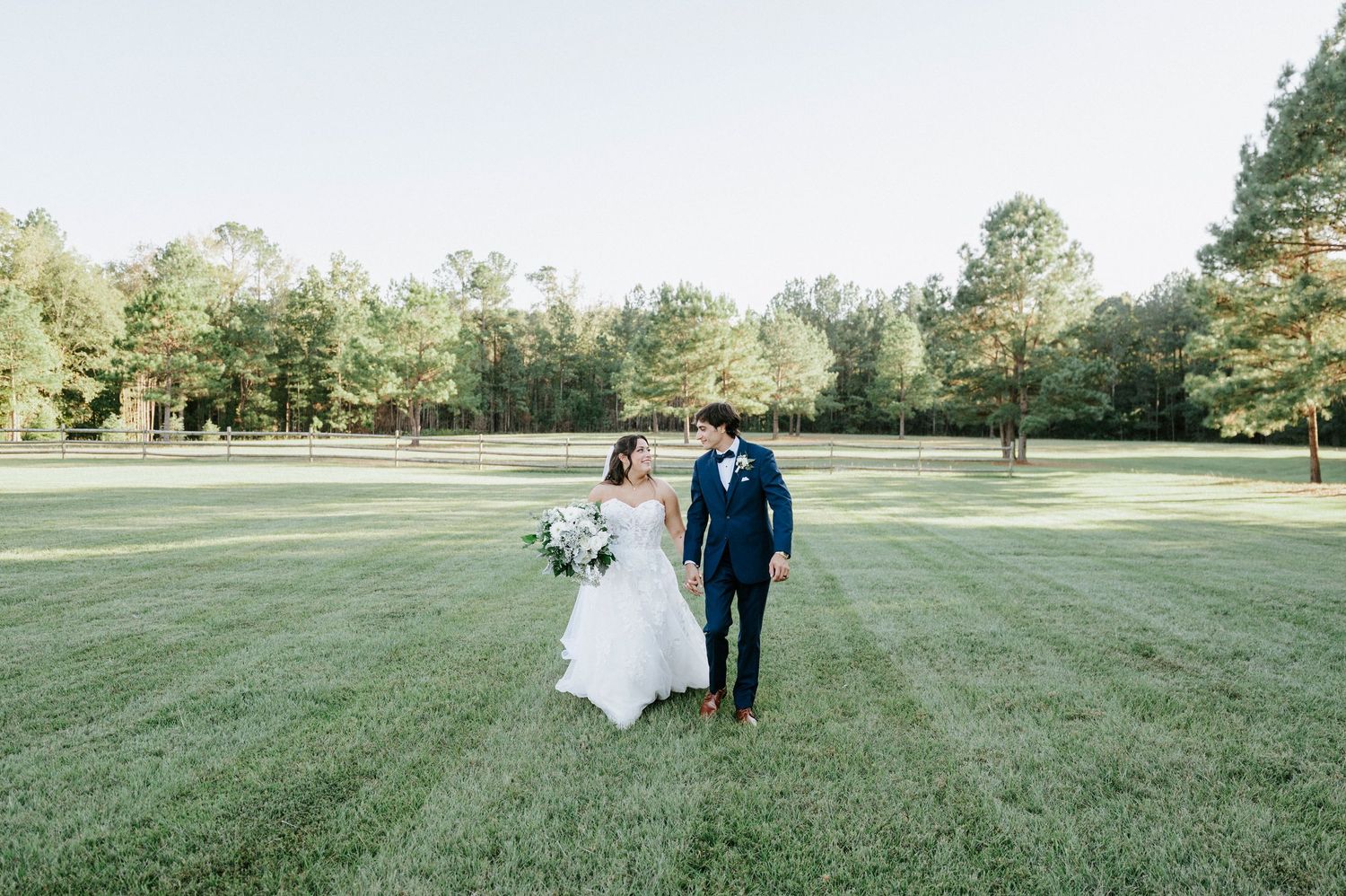 A couple in wedding attire walks together across a lush green lawn surrounded by trees on a sunny day.
