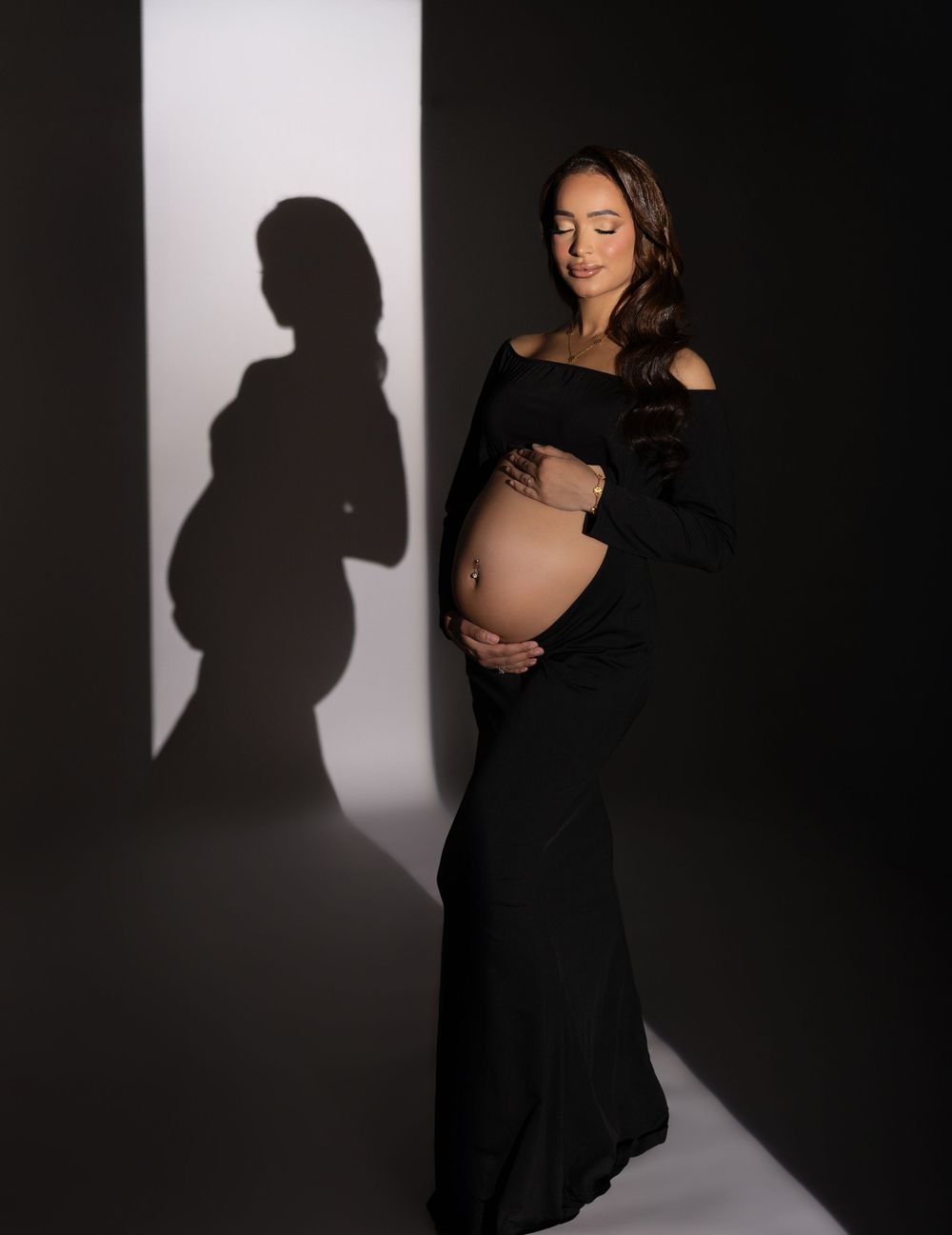 A silhouetted figure in a black dress poses against a white backdrop creating dramatic shadows in a maternity photoshoot.