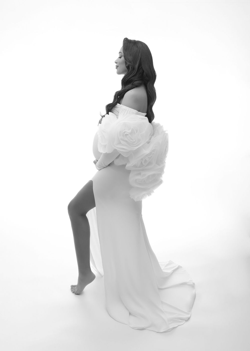 A black and white artistic maternity photo showing a woman in a flowing white dress posing elegantly against a light backdrop.