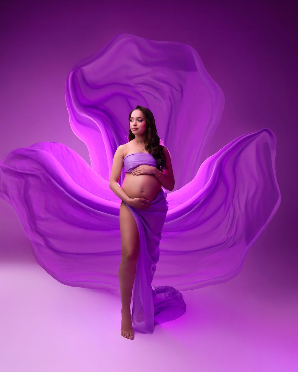 Pregnant woman in purple flowing fabric gown poses gracefully against a vibrant purple backdrop in a maternity photo shoot.