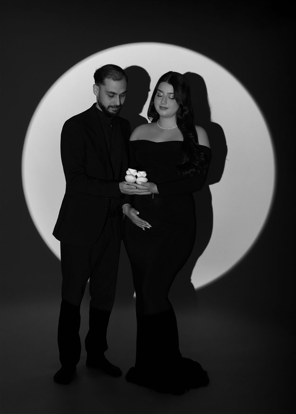 Black and white maternity photo of couple in black formal wear holding baby shoes in front of circular light backdrop.