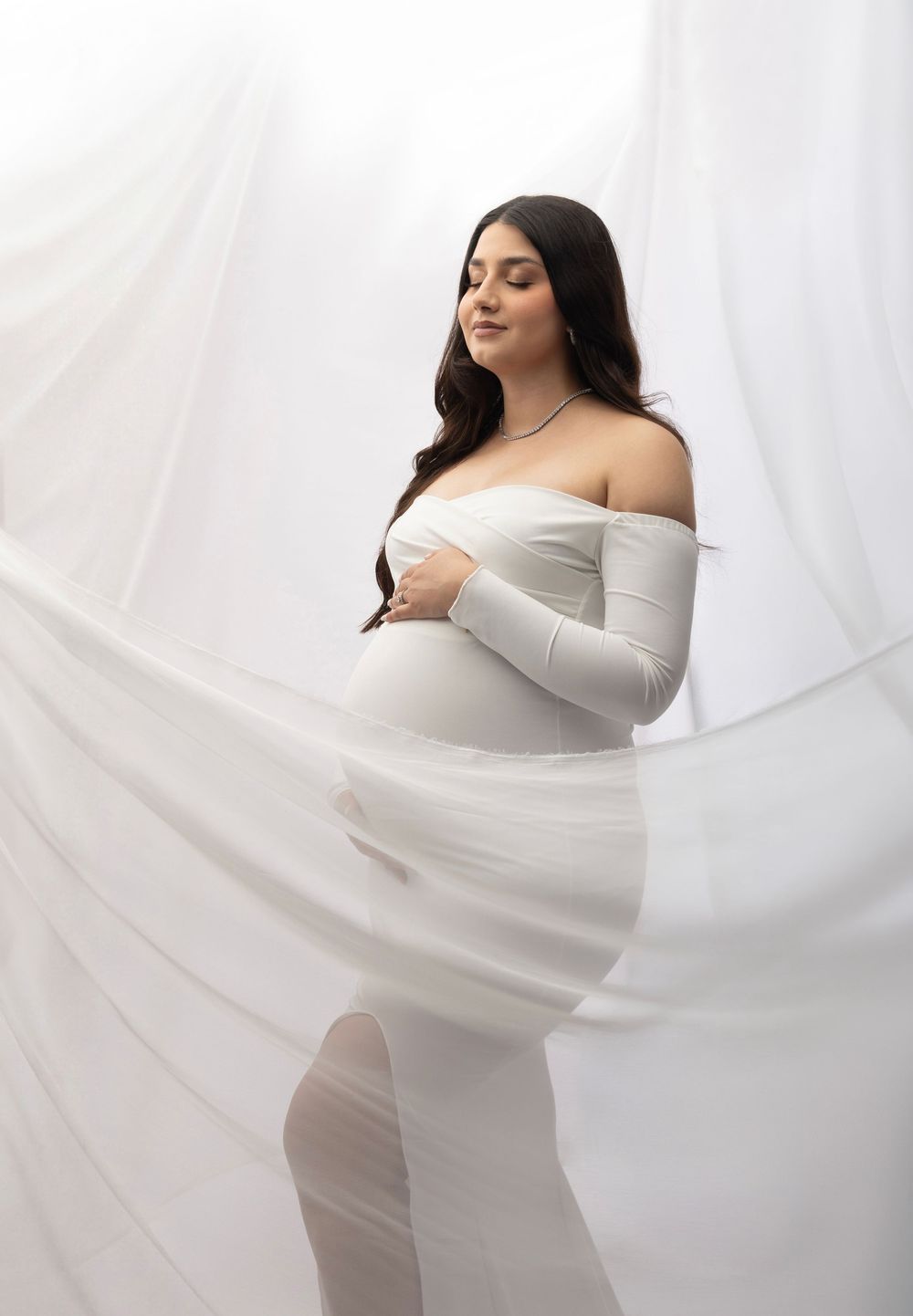 Pregnant woman in white off-shoulder gown poses gracefully with flowing fabric, maternity photography studio shoot.