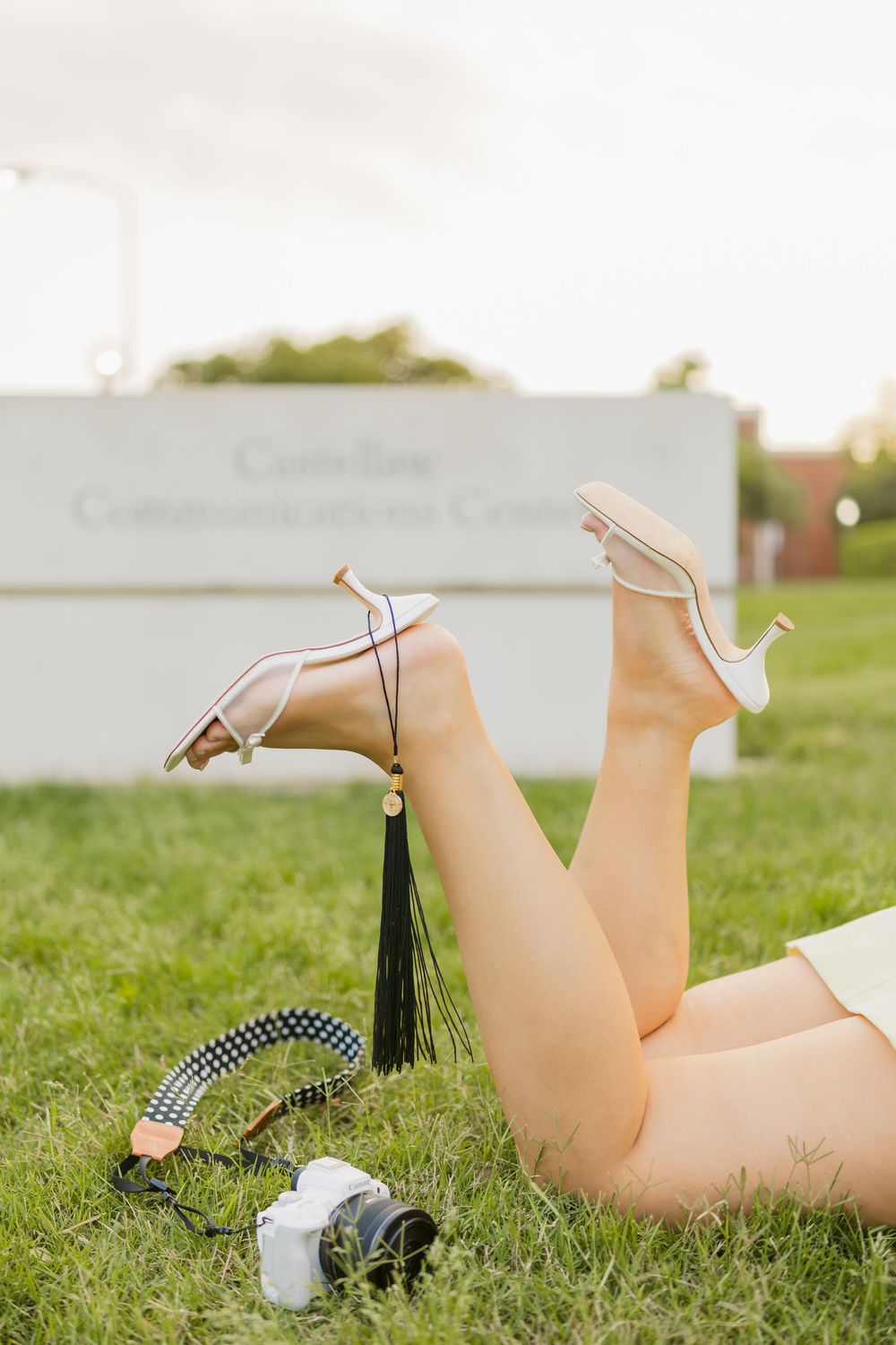 Graduate lying on grass with heels up, film camera nearby, in front of Baylor University's Castellaw Communications Center sign.