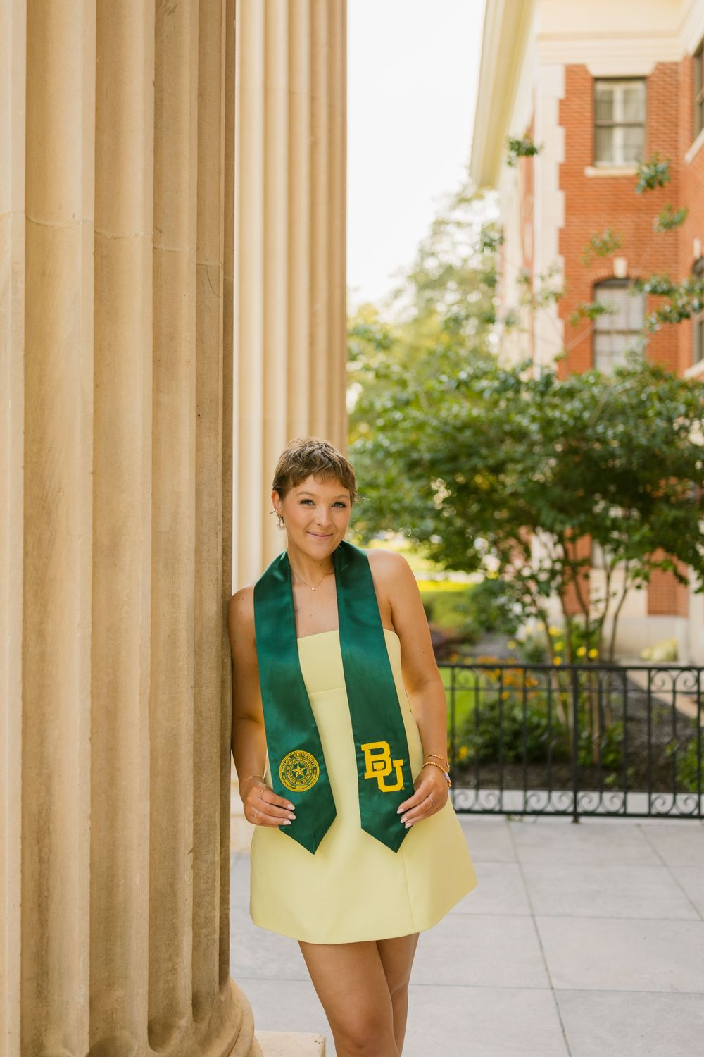 Short-haired female Baylor University graduate in yellow dress and green BU stole leaning against campus column.