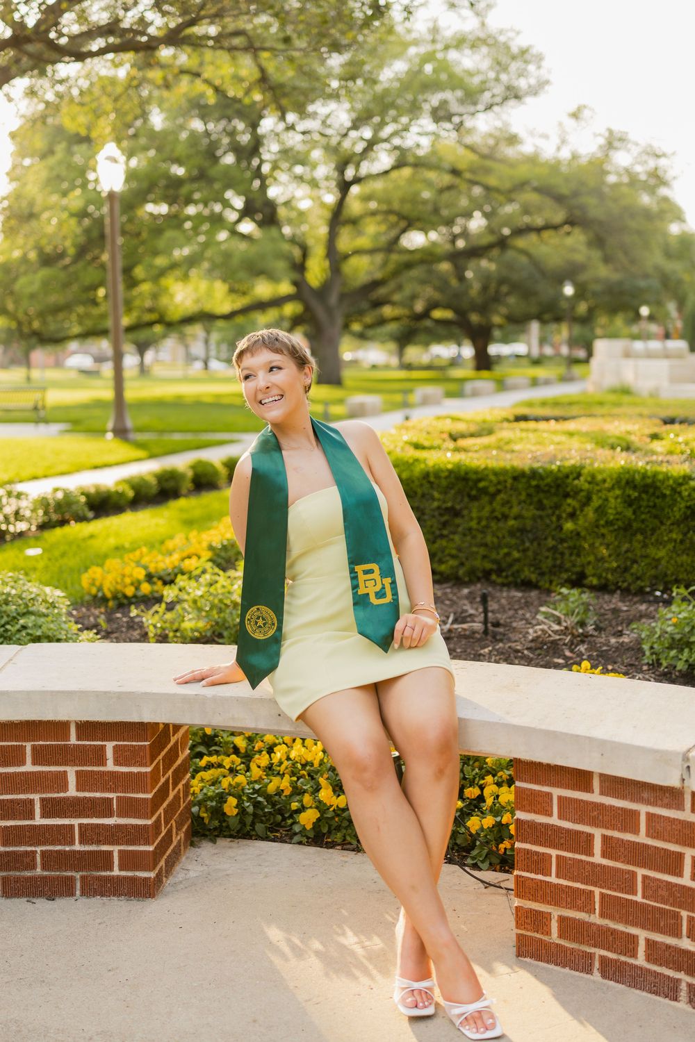 Baylor University graduate smiling in a yellow dress with a green BU stole, seated on a brick wall surrounded by flowers.