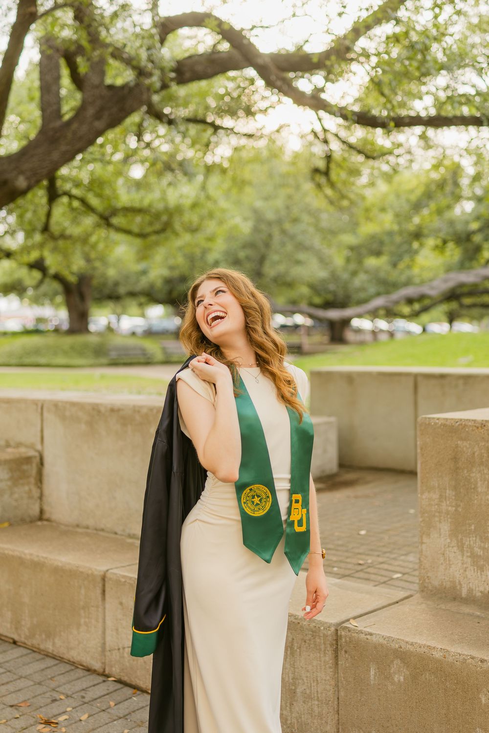Joyful female Baylor graduate in cream dress holding graduation gown and green BU stole outdoors.