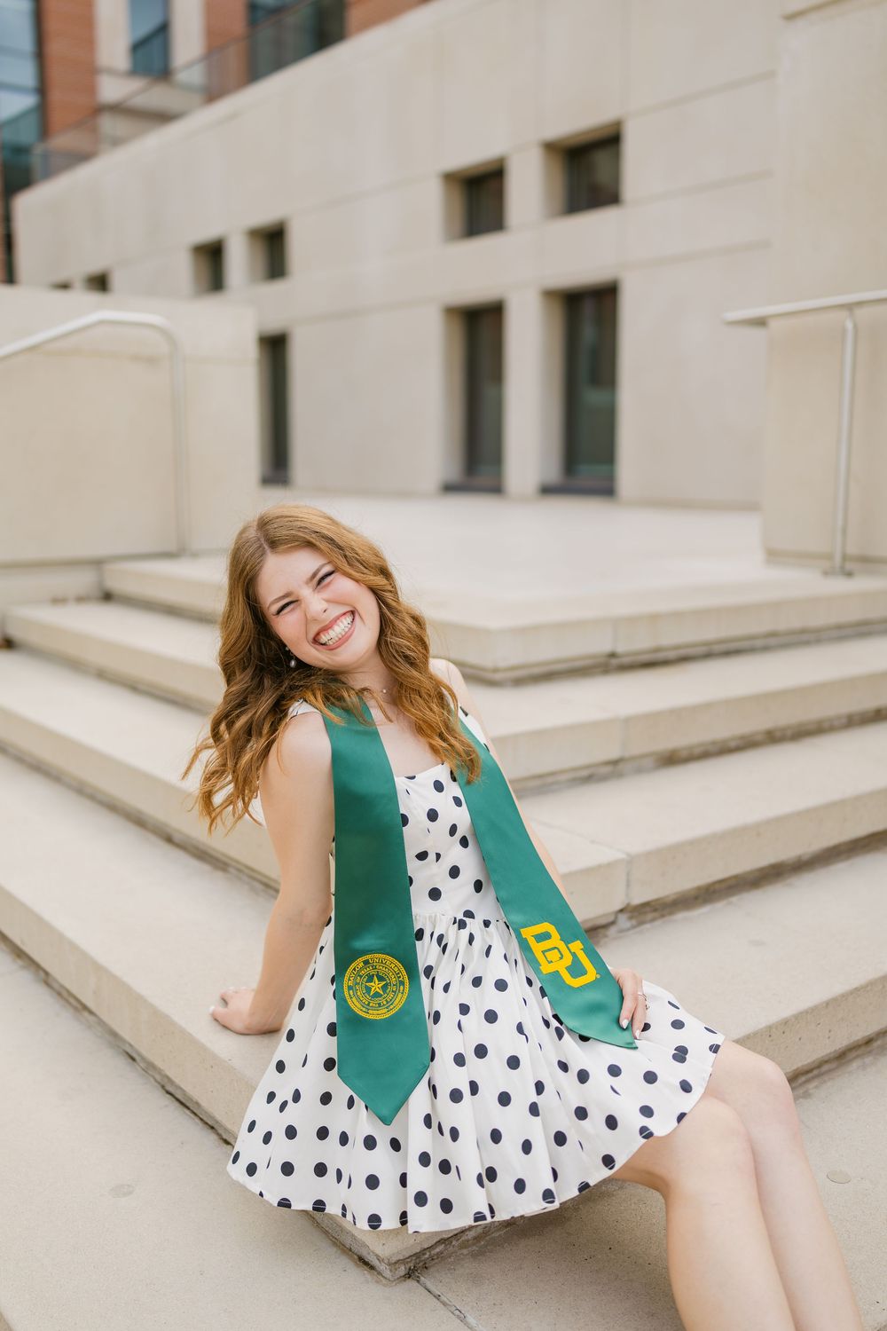 Smiling redhead graduate in polka dot dress and green Baylor University stole sitting on campus steps.