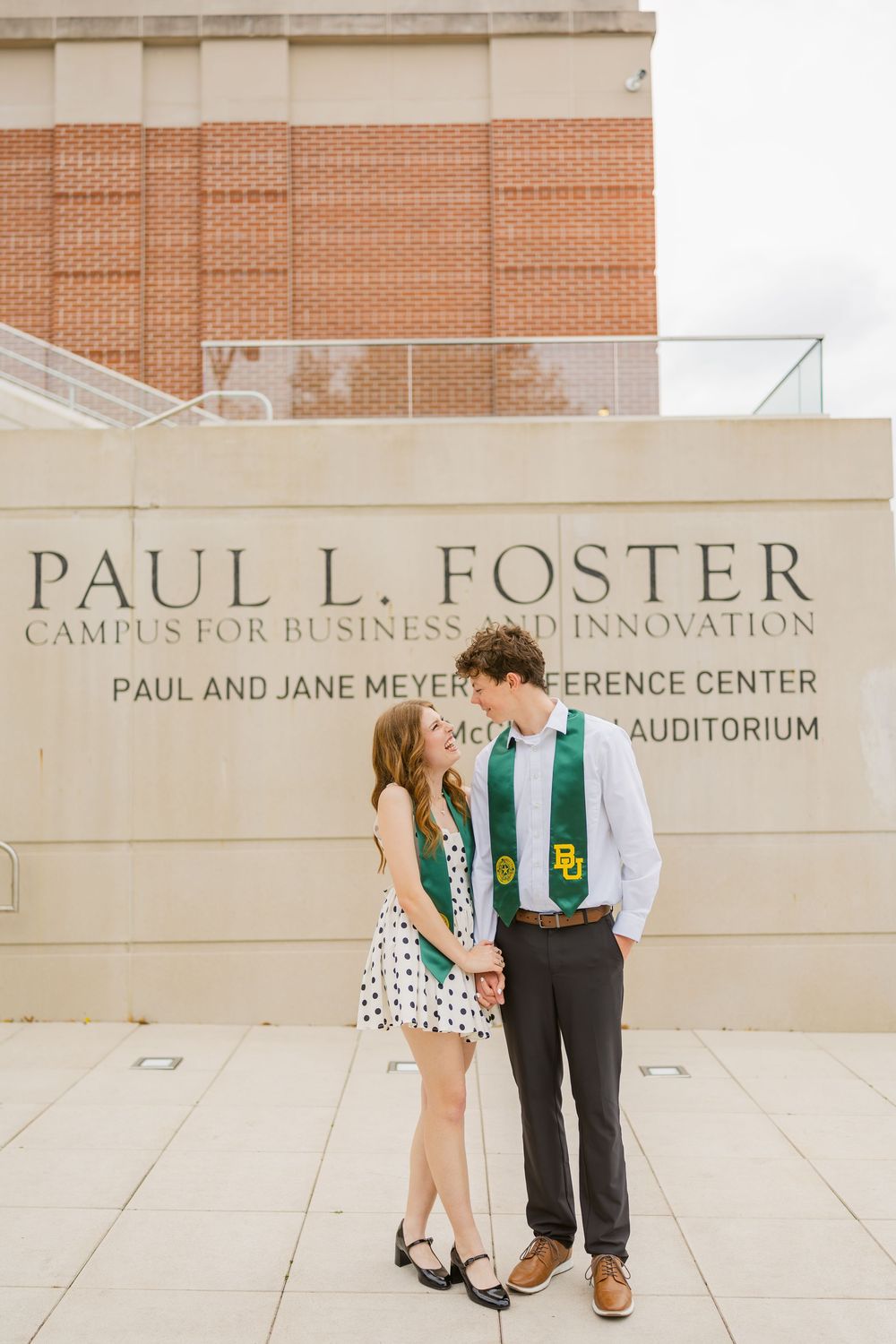 Two Baylor University graduates in green stoles standing together at Paul L. Foster Campus for Business.