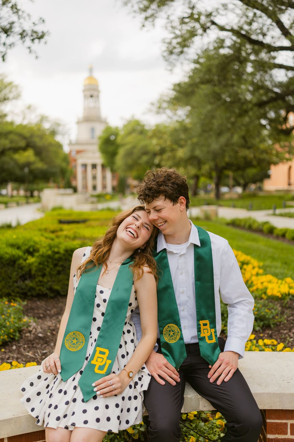 Two laughing Baylor graduates in green BU stoles sitting outdoors near iconic campus bell tower.