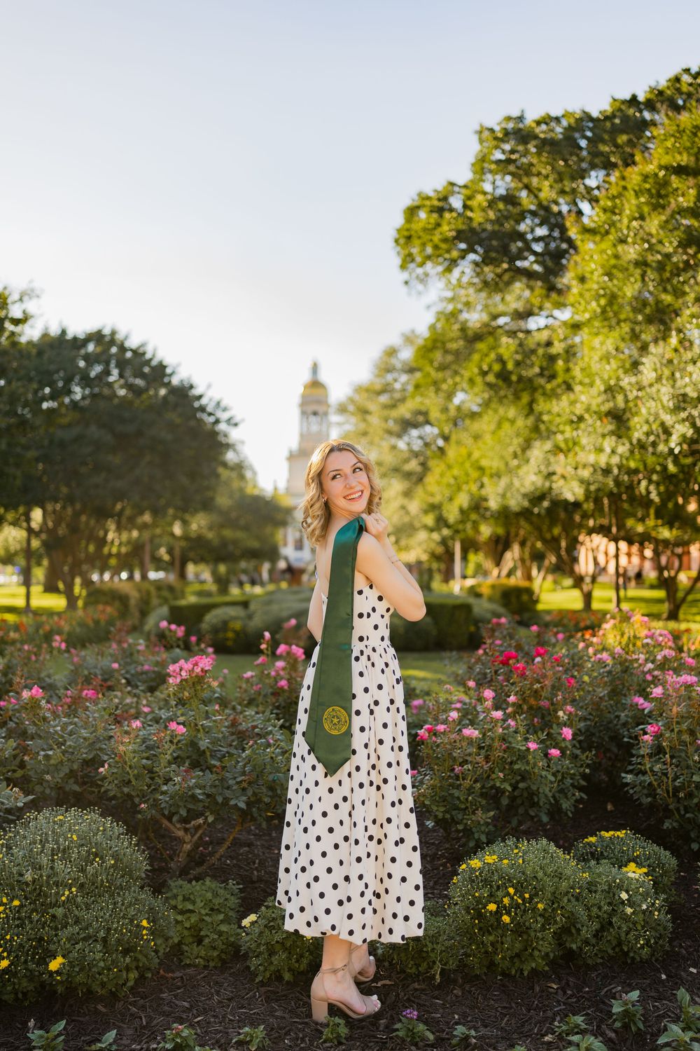 Smiling blonde Baylor graduate in polka dot dress standing in campus rose garden with bell tower.