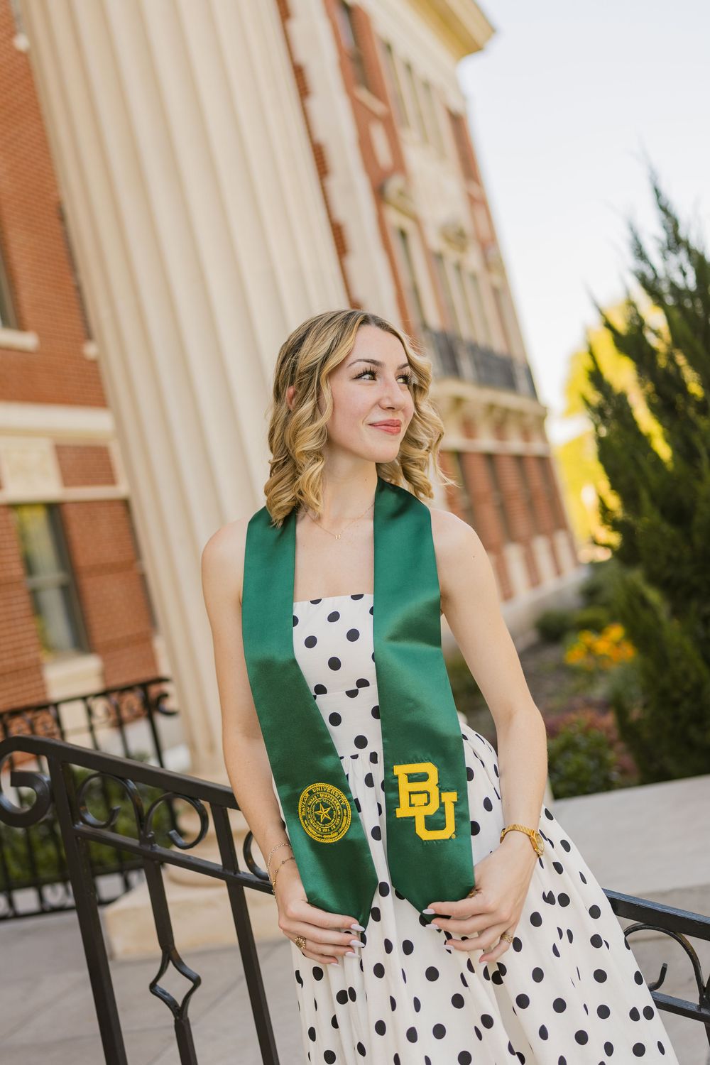 Blonde female graduate in polka dot dress wearing green Baylor University stole on campus steps.