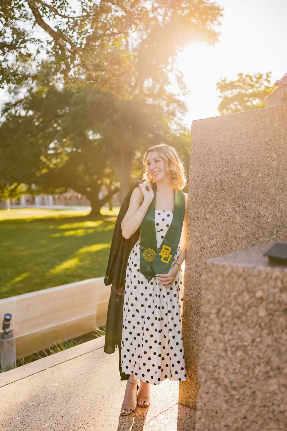 Blonde female Baylor graduate in a polka dot dress, posing in golden sunlight, holding a graduation gown.