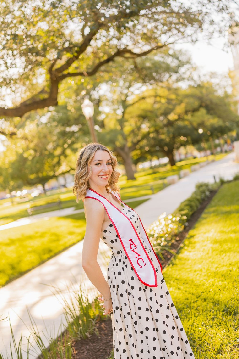 Smiling blonde graduate in a polka dot dress wearing a red and white sash walking on a sunny campus path.