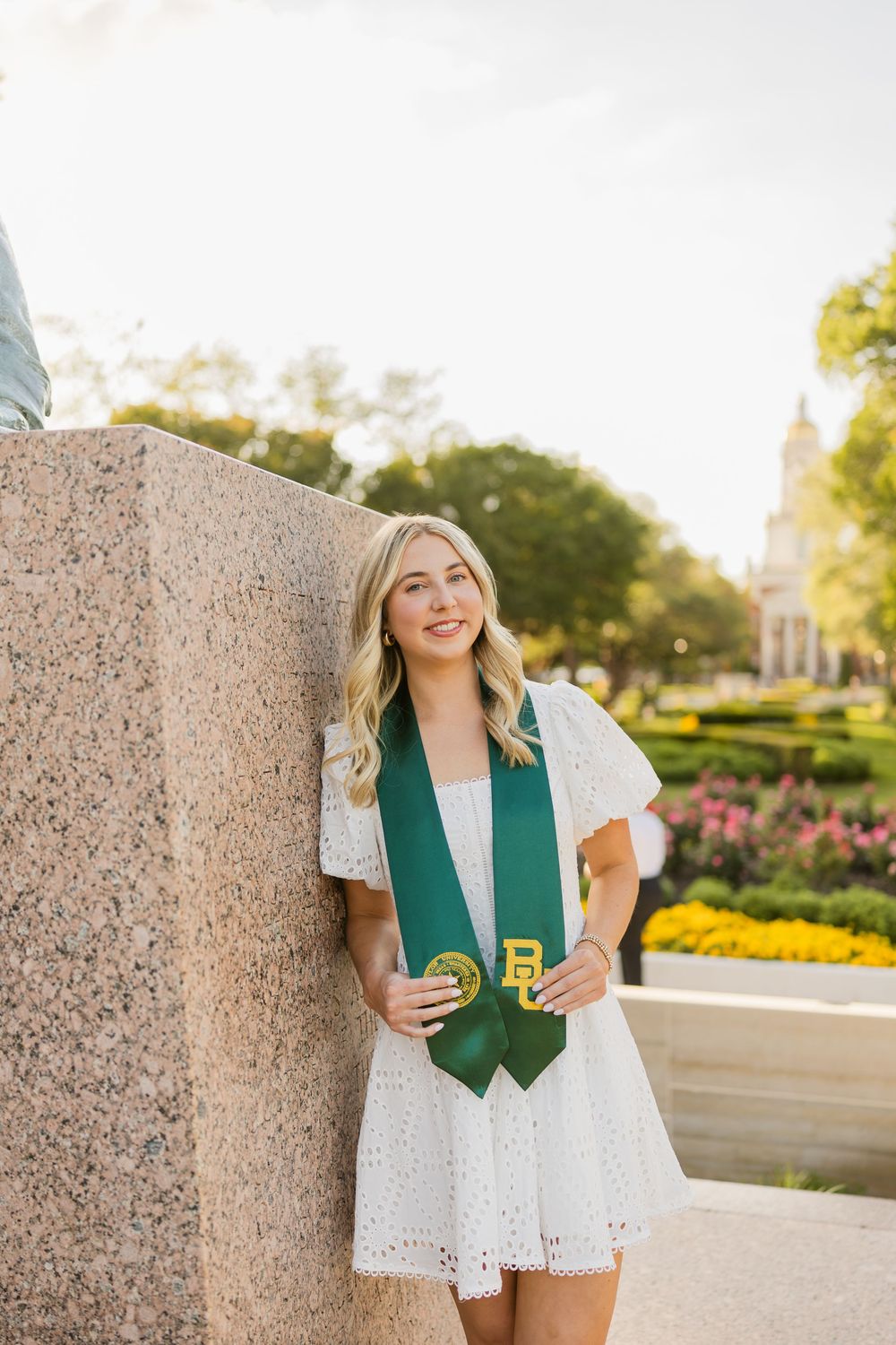 Blonde woman in a white dress, wearing a Baylor University graduation stole, leaning against a stone monument.
