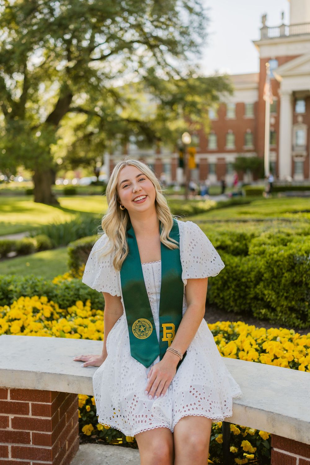 Smiling blonde woman in white eyelet dress with green Baylor graduation stole seated by yellow flowers.