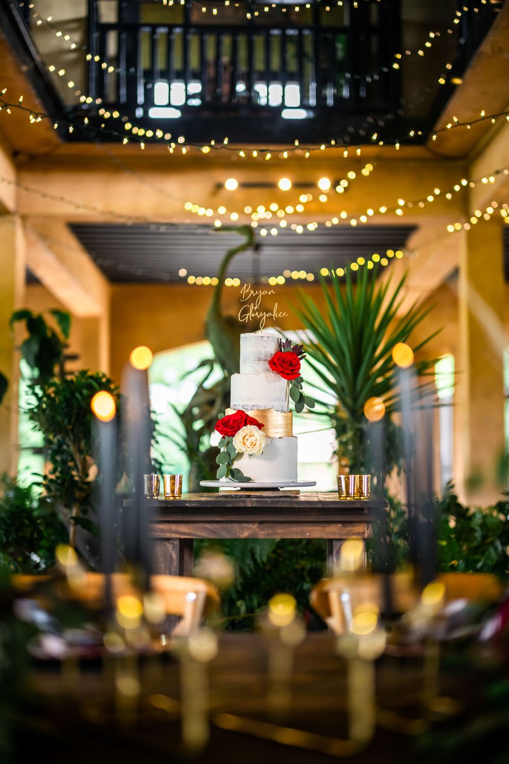 Elegant white wedding cake displayed on wooden table with string lights and tropical plants in the background.