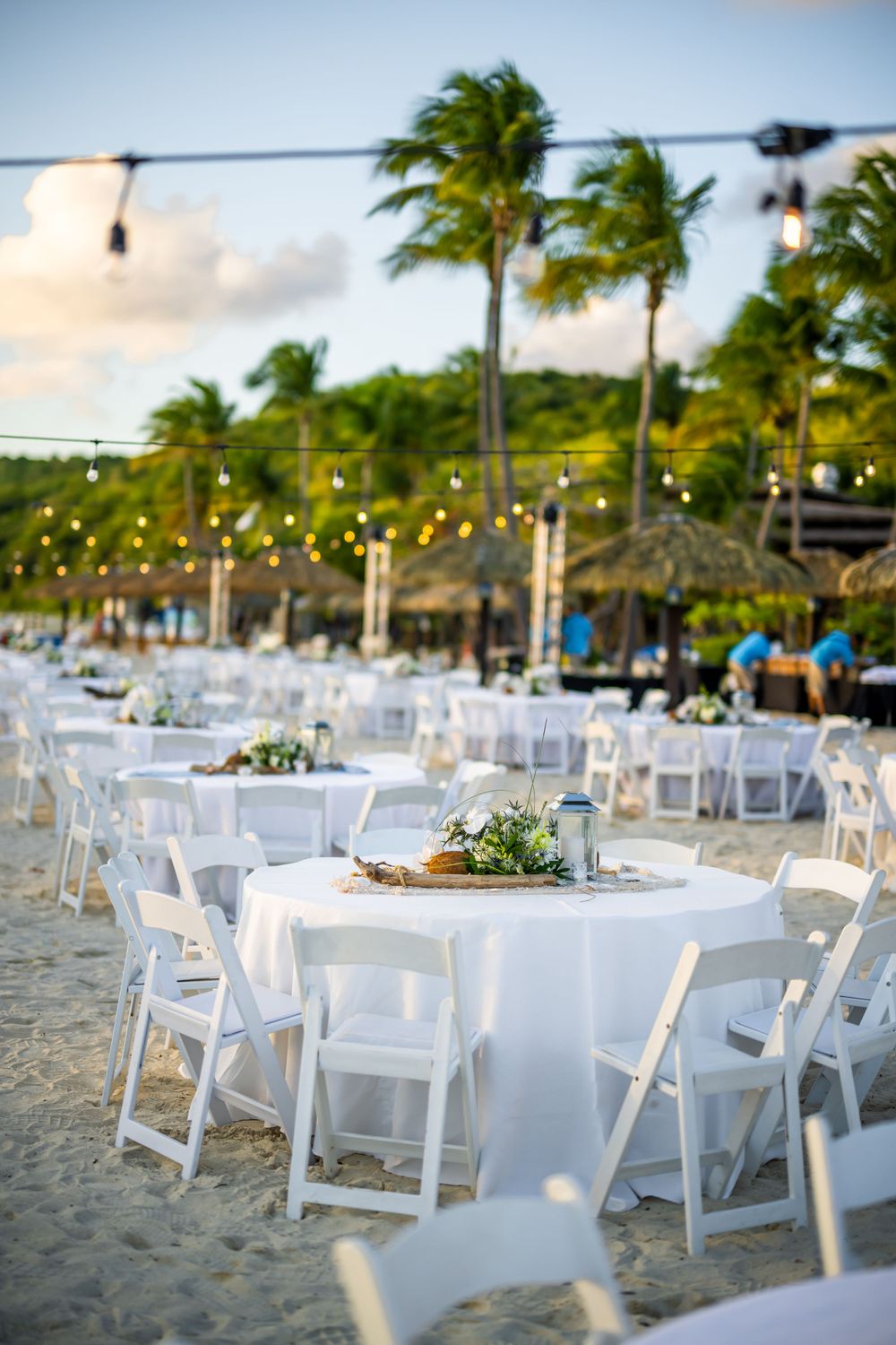 Beach wedding reception setup with white tables and string lights under palm trees at sunset.