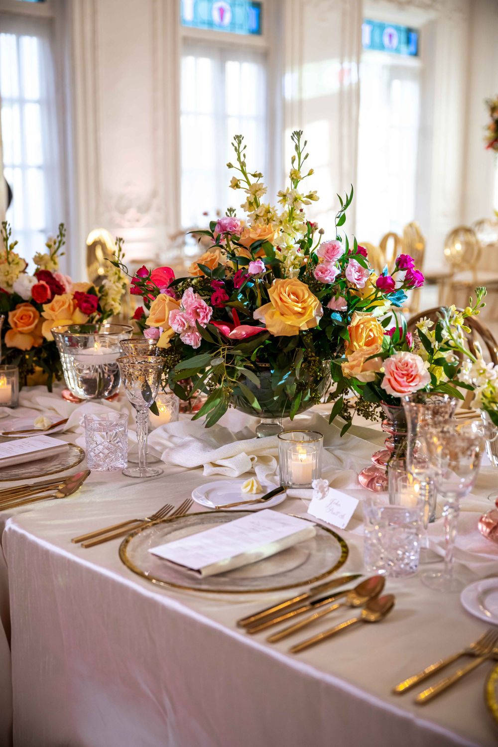 Elegant table setting with gold flatware and vibrant floral centerpiece featuring roses and mixed blooms.