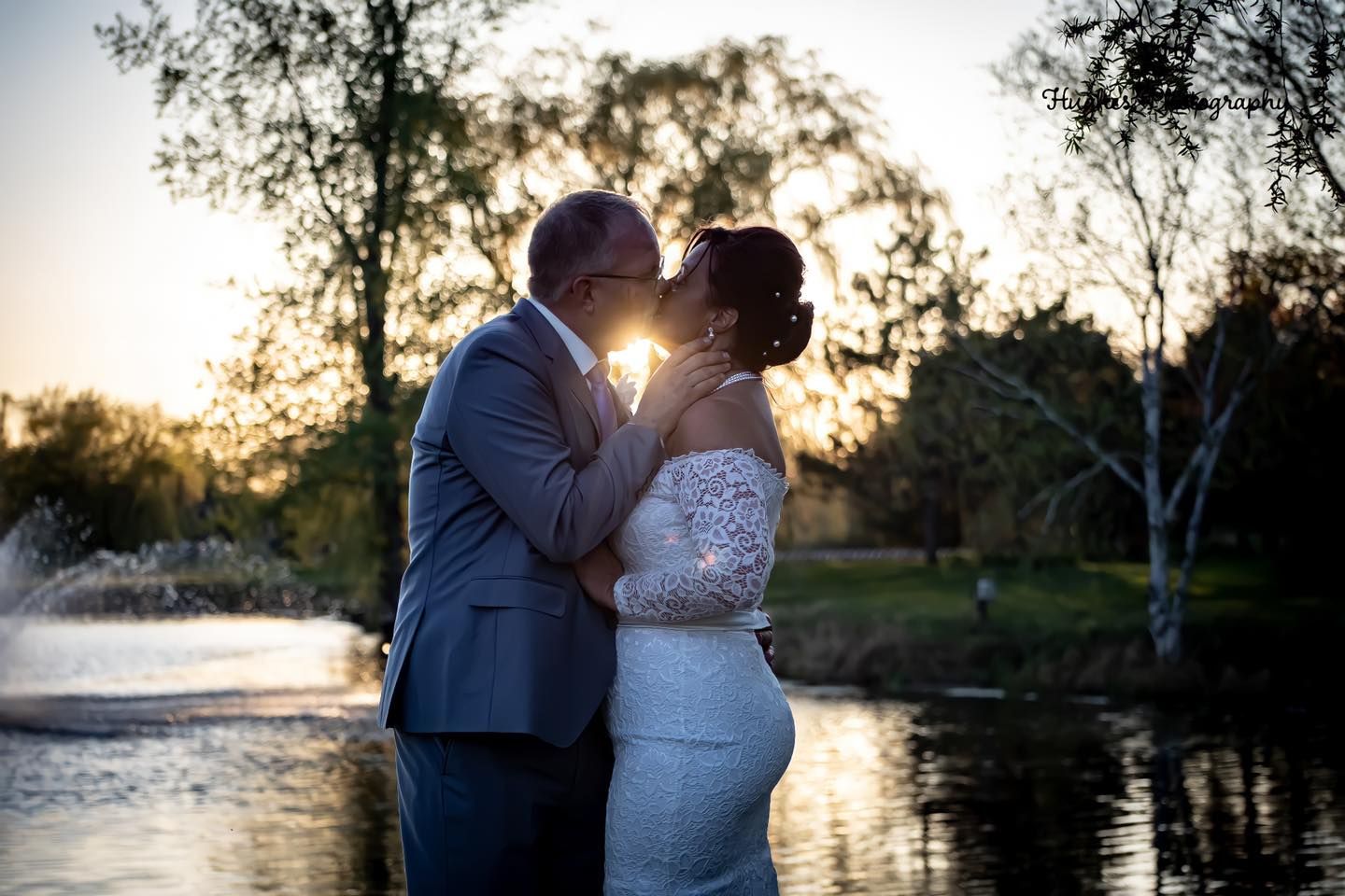 A silhouetted couple shares a kiss at sunset by a tranquil lake with trees in the background.