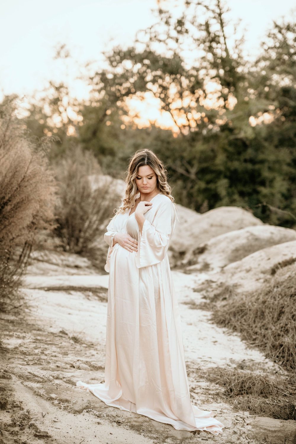 Figure in white flowing dress poses gracefully on a sandy path at sunset.
