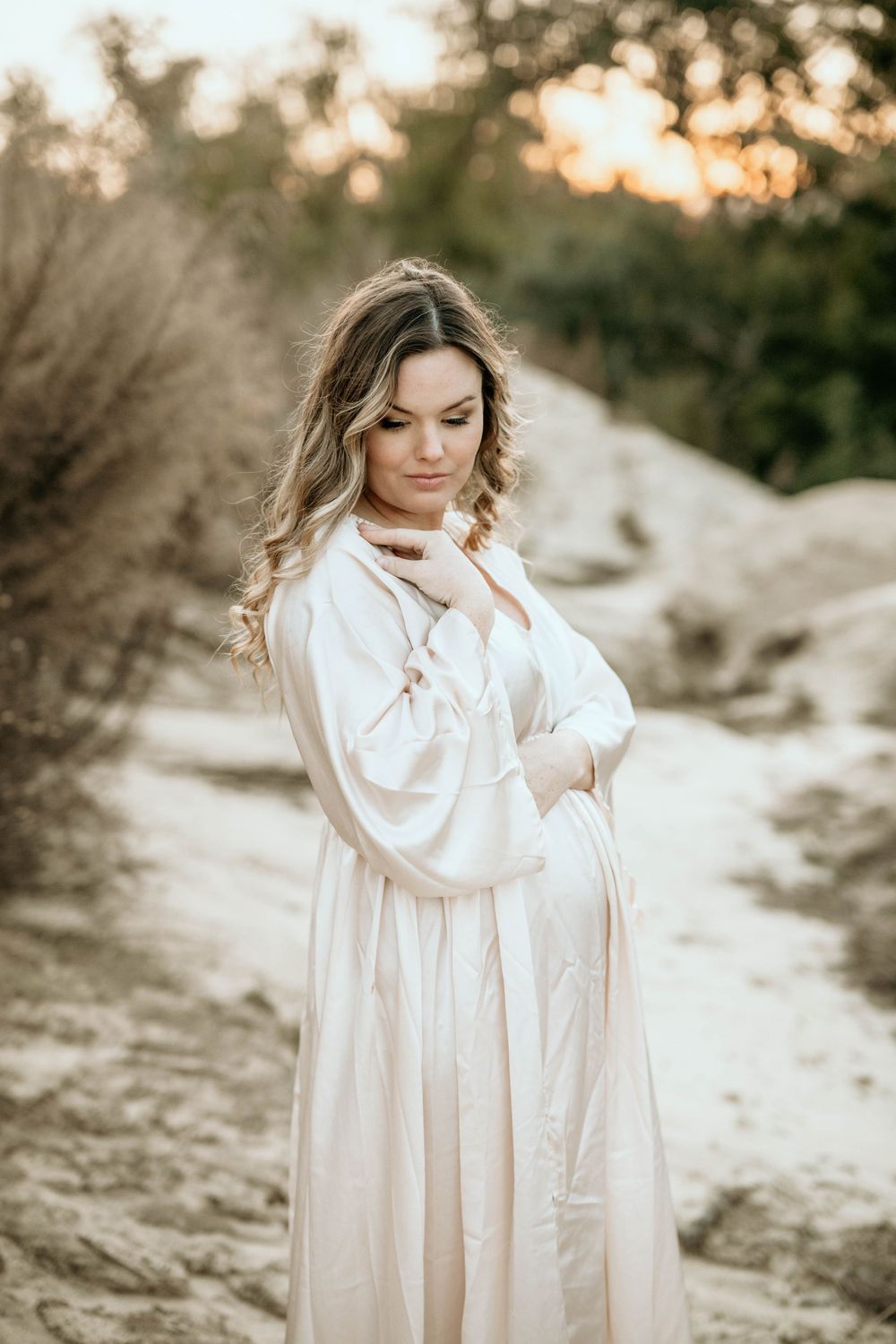 Person in elegant white dress poses thoughtfully against a sunset desert backdrop.