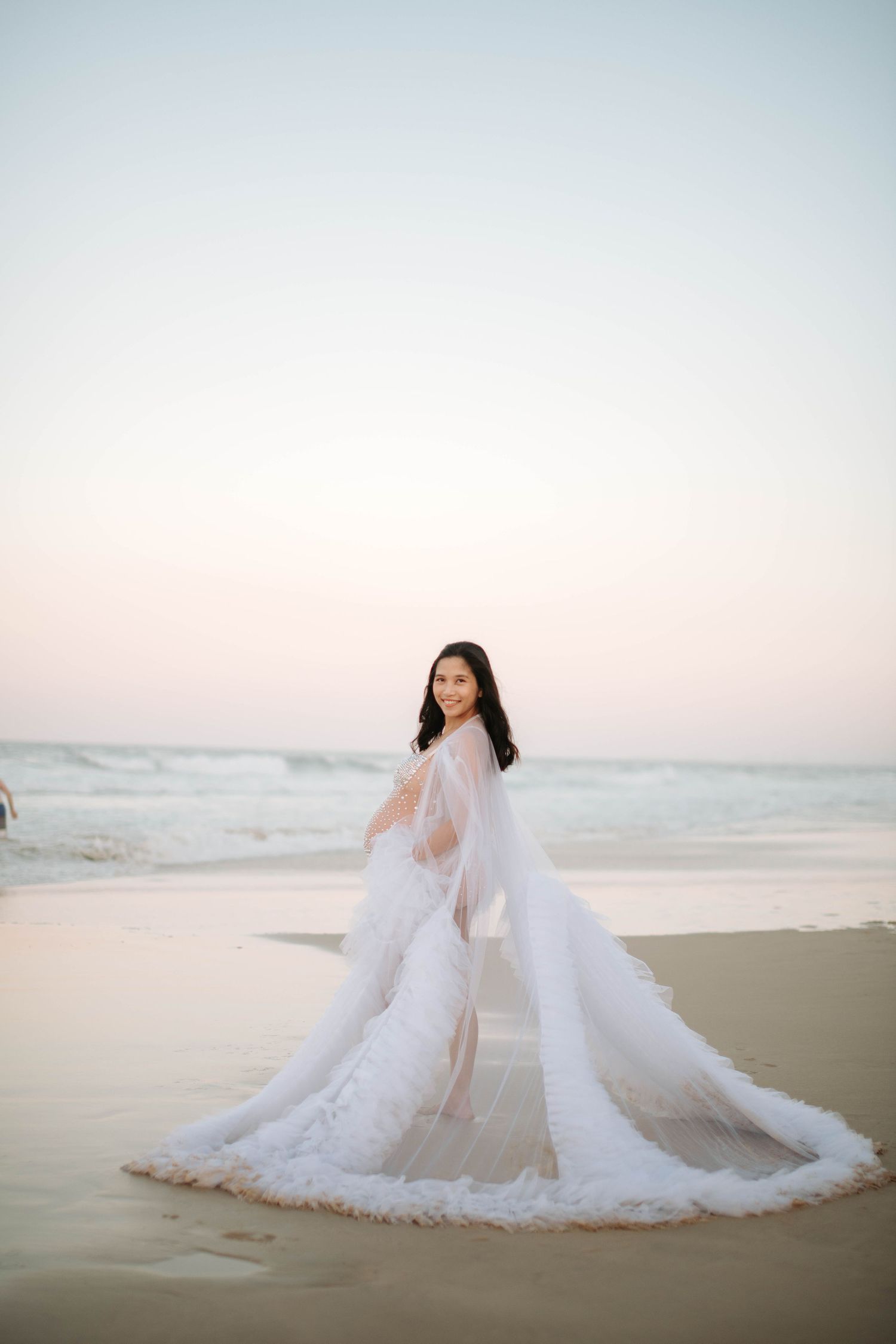 Elegant maternity photo of a woman in flowing white dress on beach at sunset.