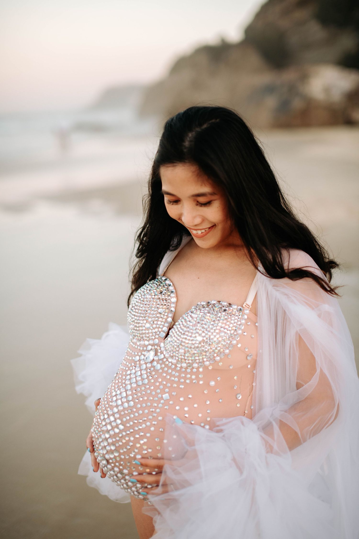 A maternity portrait featuring a sparkly sheer dress and flowing white cape on a cloudy beach.
