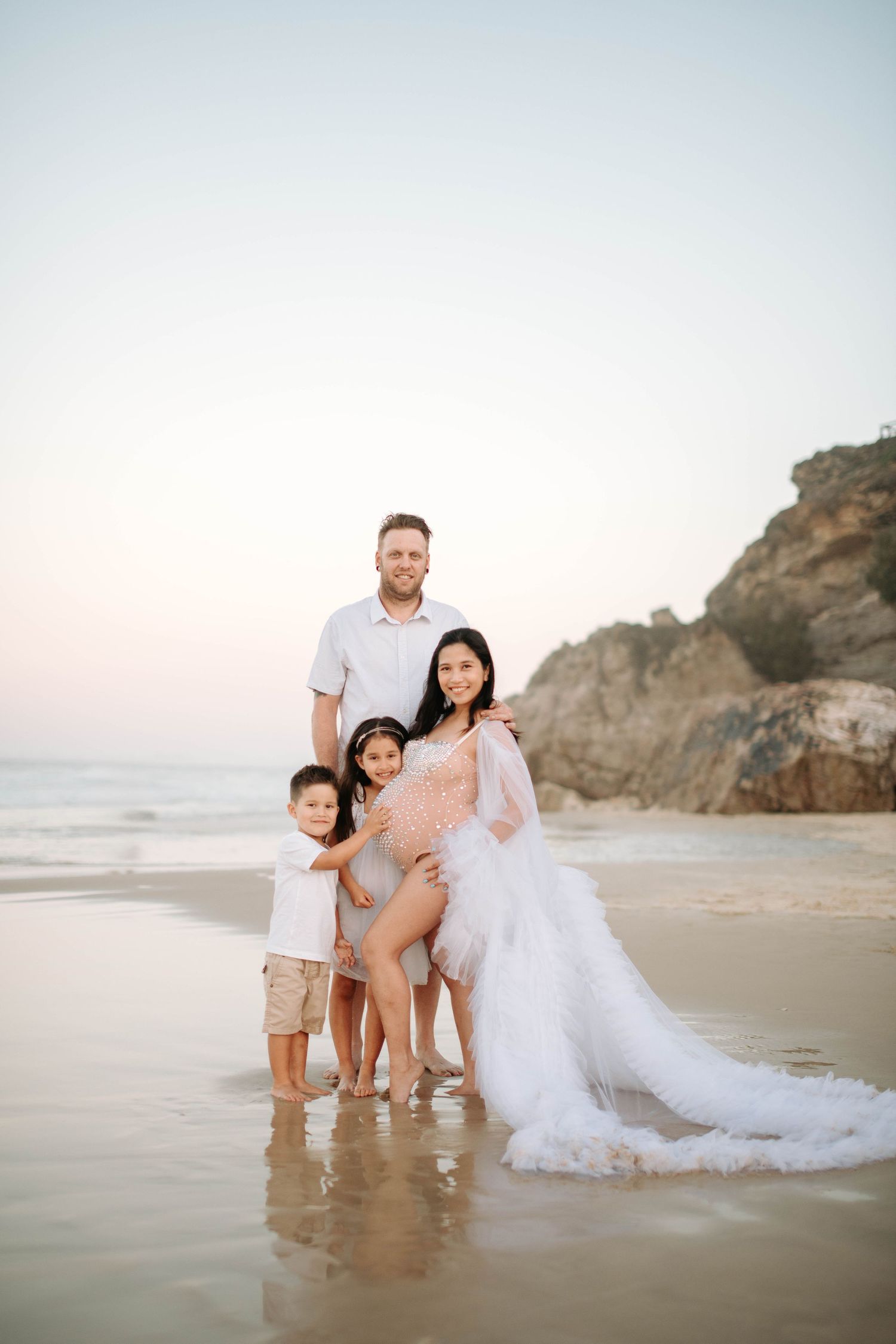 Beach family portrait with flowing white dress against rocky coastal backdrop.