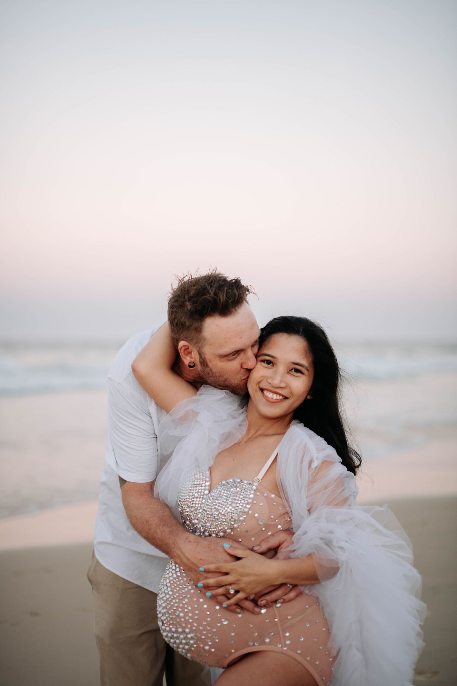 A romantic beachside maternity photo with a sparkling sheer dress and flowing white cape at sunset.