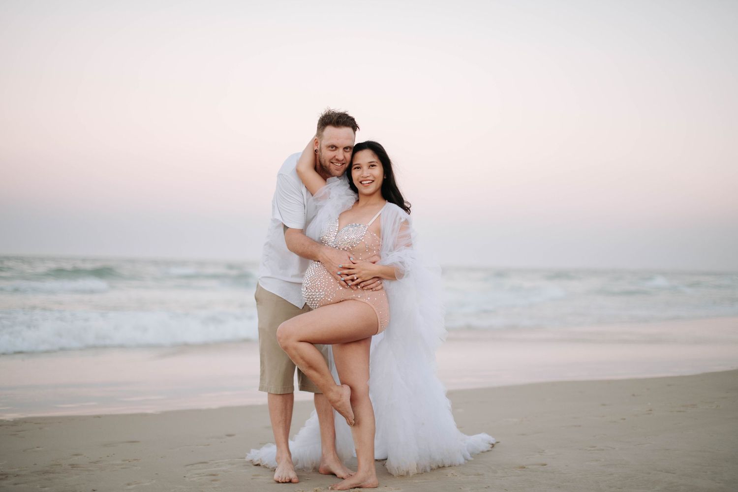 A couple poses together on a beach at sunset wearing white and beige casual attire.