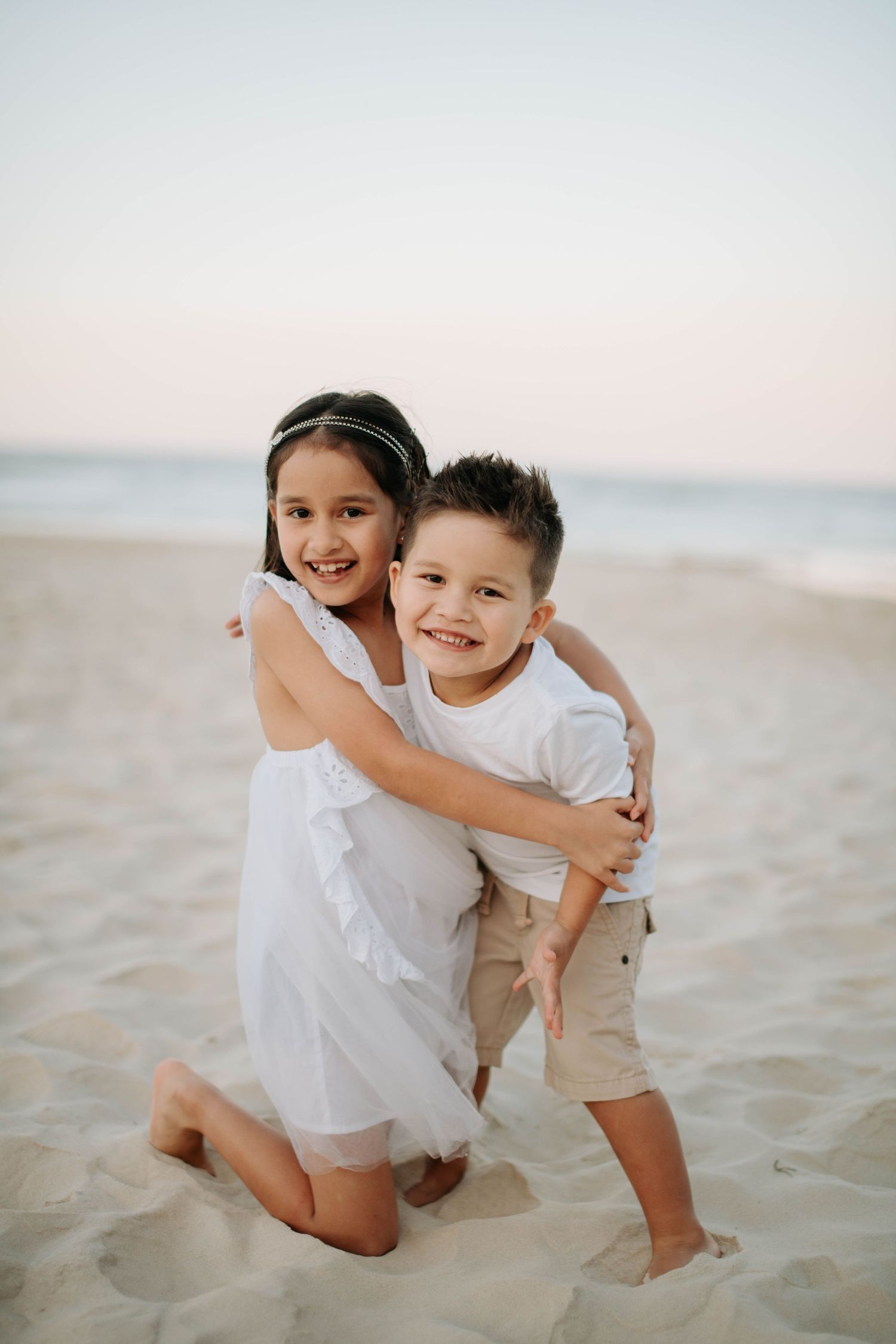 Two children in white outfits embrace and smile while playing on a sandy beach.