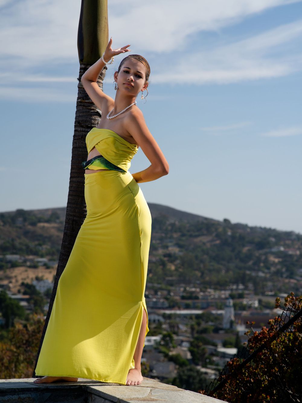 Model in flowing yellow gown poses against cityscape view from hilltop location.