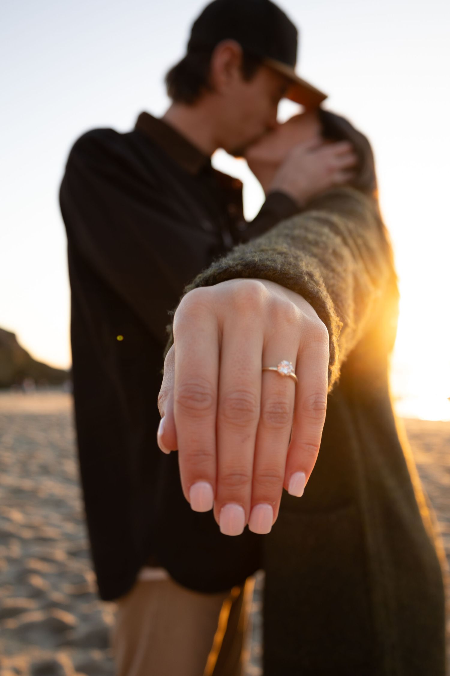 A newly engaged couple embrace each other in a kiss while the bride to be shows off her engagement ring on a beach photoshoot. A sun flare