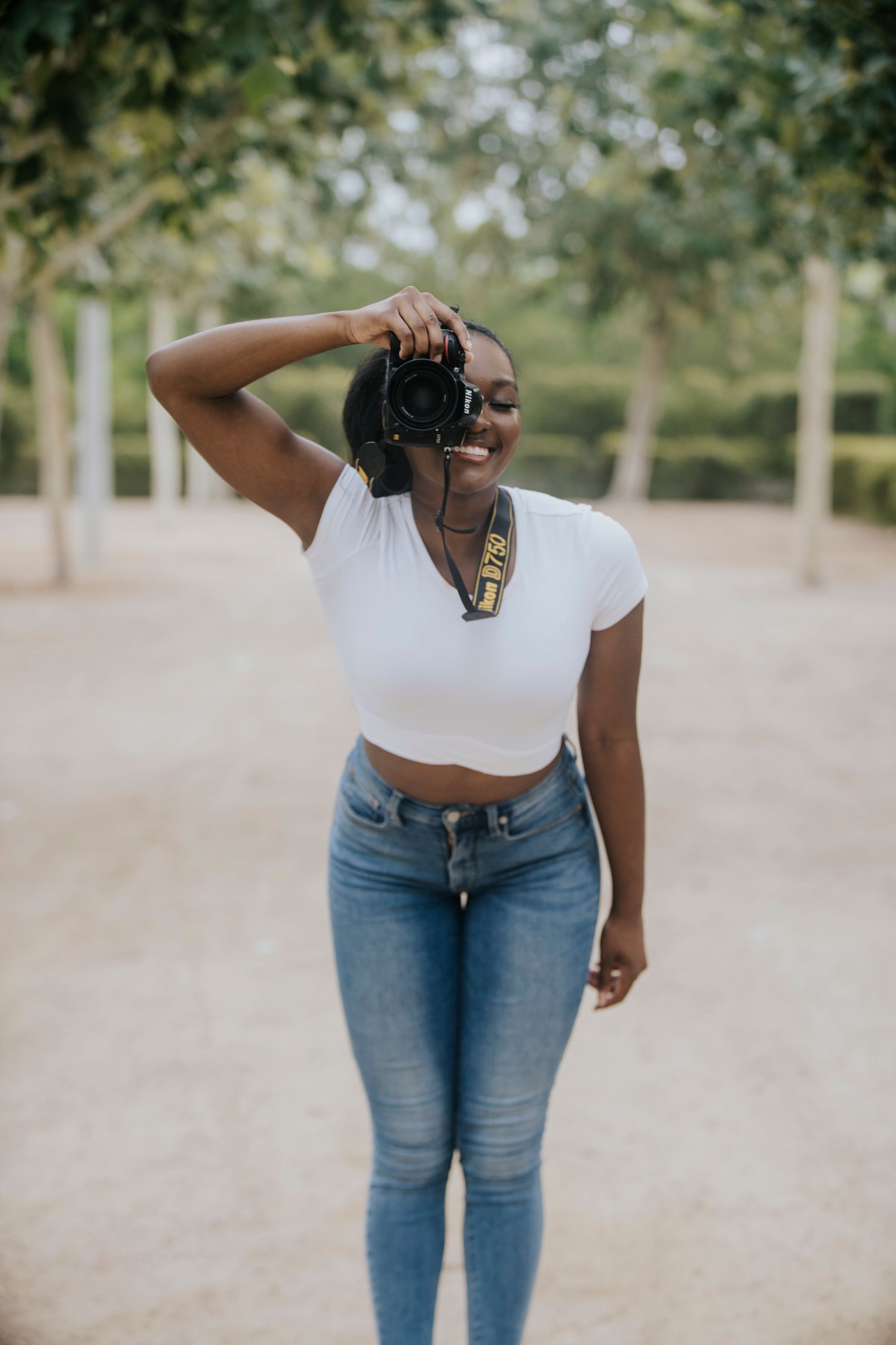 Photographer in white crop top and jeans takes pictures in a tree-lined pathway in Buffalo Bayou Park