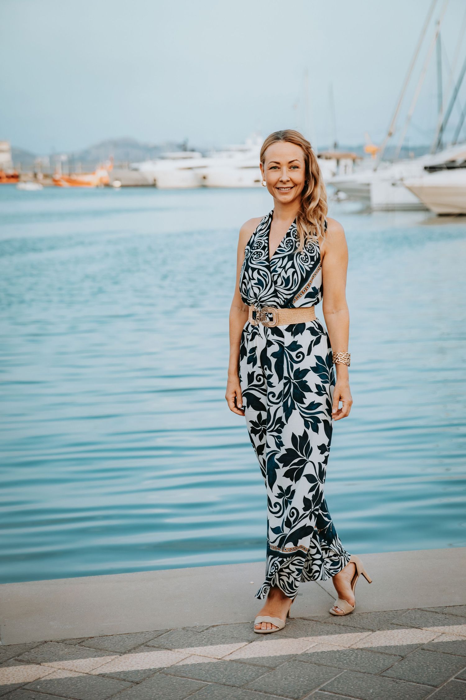 Full length portrait of a woman wearing a floral summer dress outfit walking in a maritime environment with boats in the background.