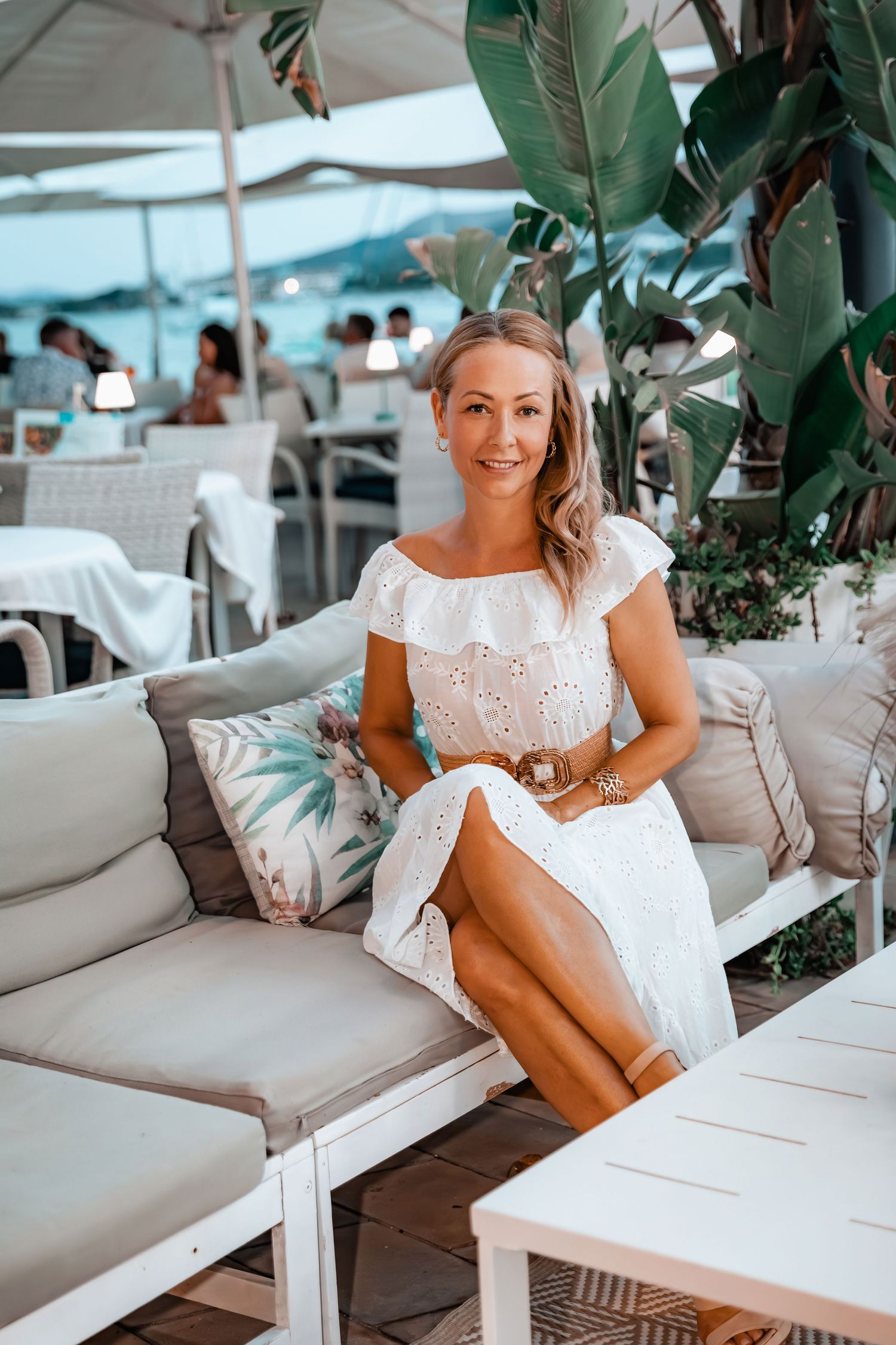 Relaxed woman smiling sitting on a white lounge sofa in white summer-dress at beach club NU Mallorca, Playa de Muro.