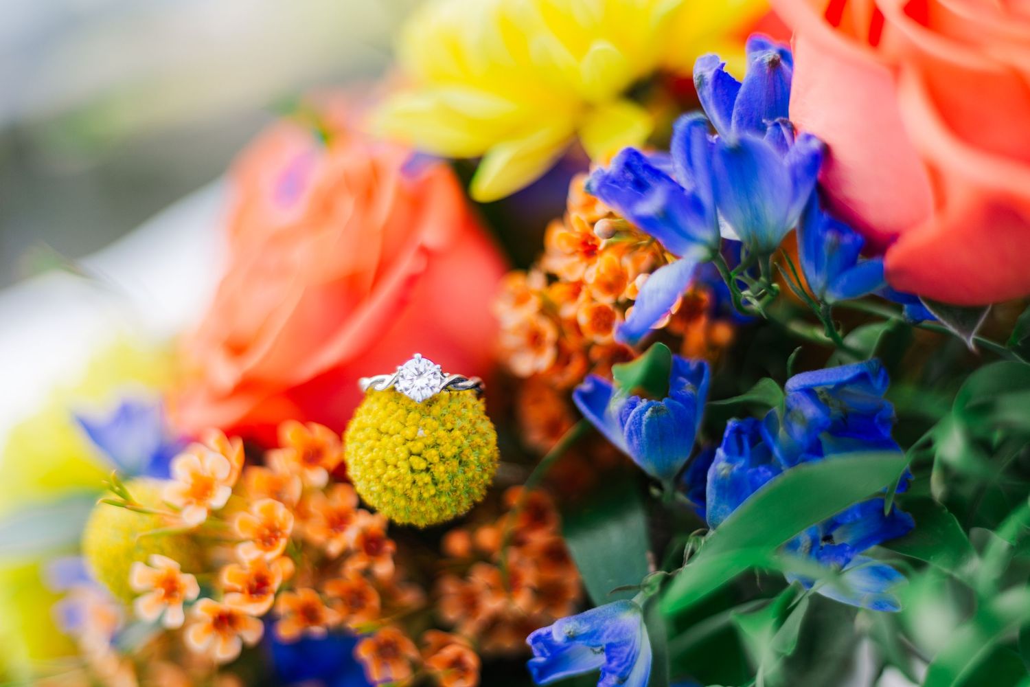 Vibrant wedding bouquet featuring orange roses, yellow craspedia, blue delphinium and a sparkling engagement ring.