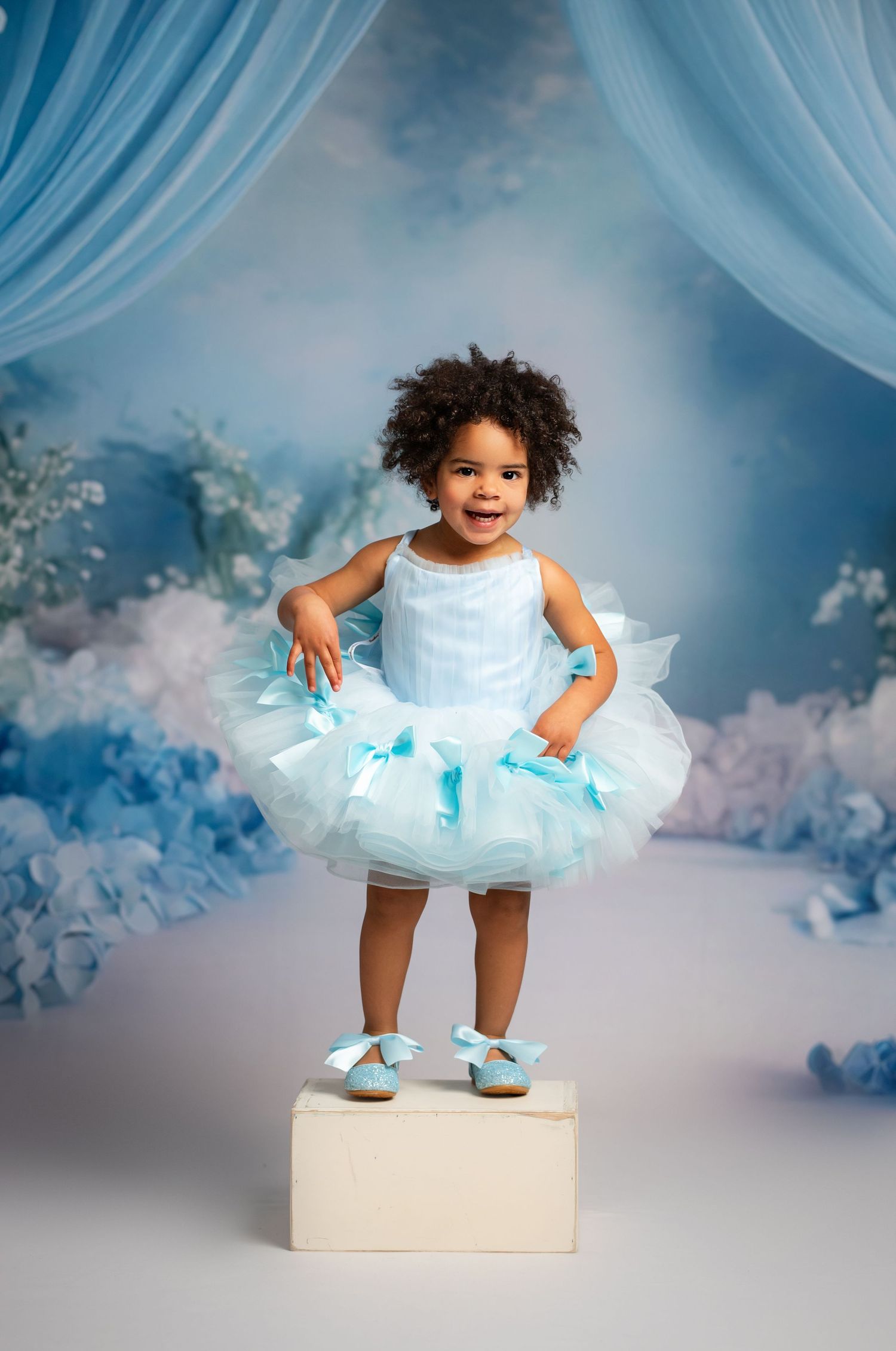 Young performer in light blue tutu dress and ballet shoes stands on white block against dreamy cloudy backdrop.