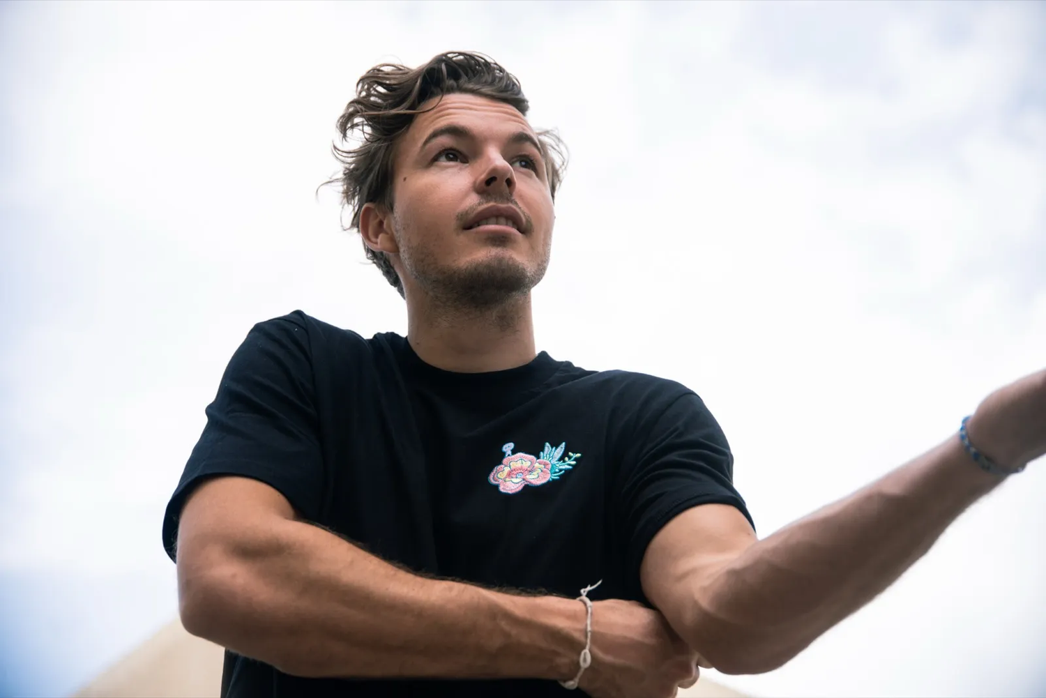Un jeune homme se tient les bras croisés, regardant au loin, capté sous un ciel lumineux.