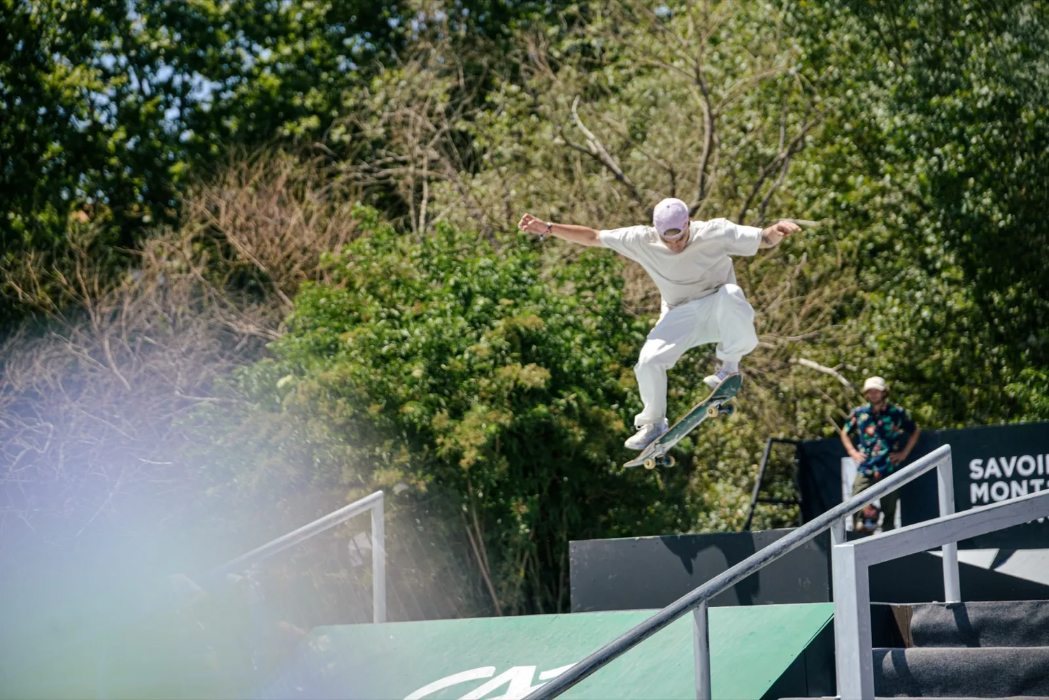 Un skateur réalise un saut technique sur une rampe en pleine compétition, entouré d’arbres et sous un ciel dégagé.