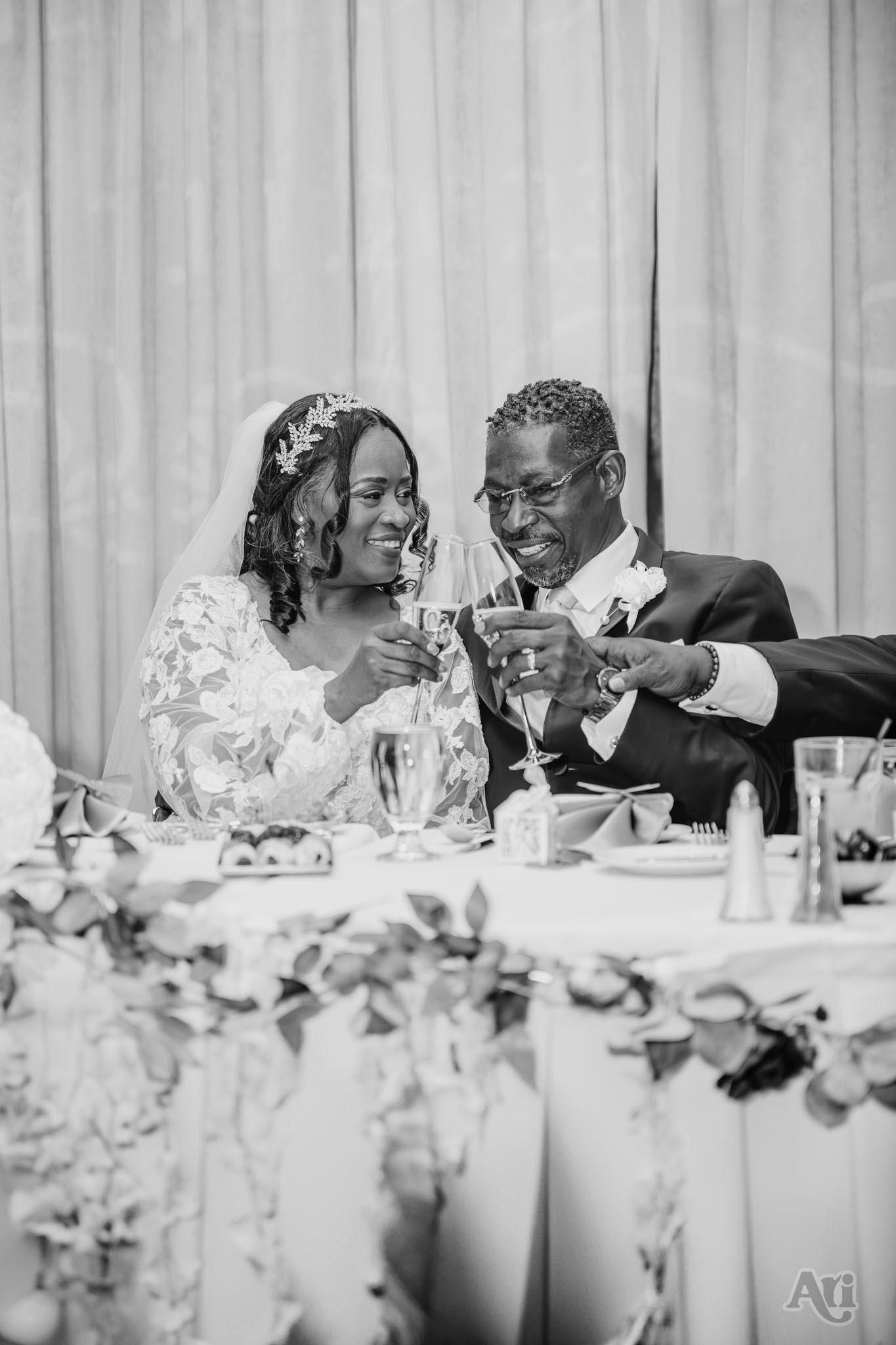 Black and white wedding reception photo of newlyweds sharing drinks at decorated table.