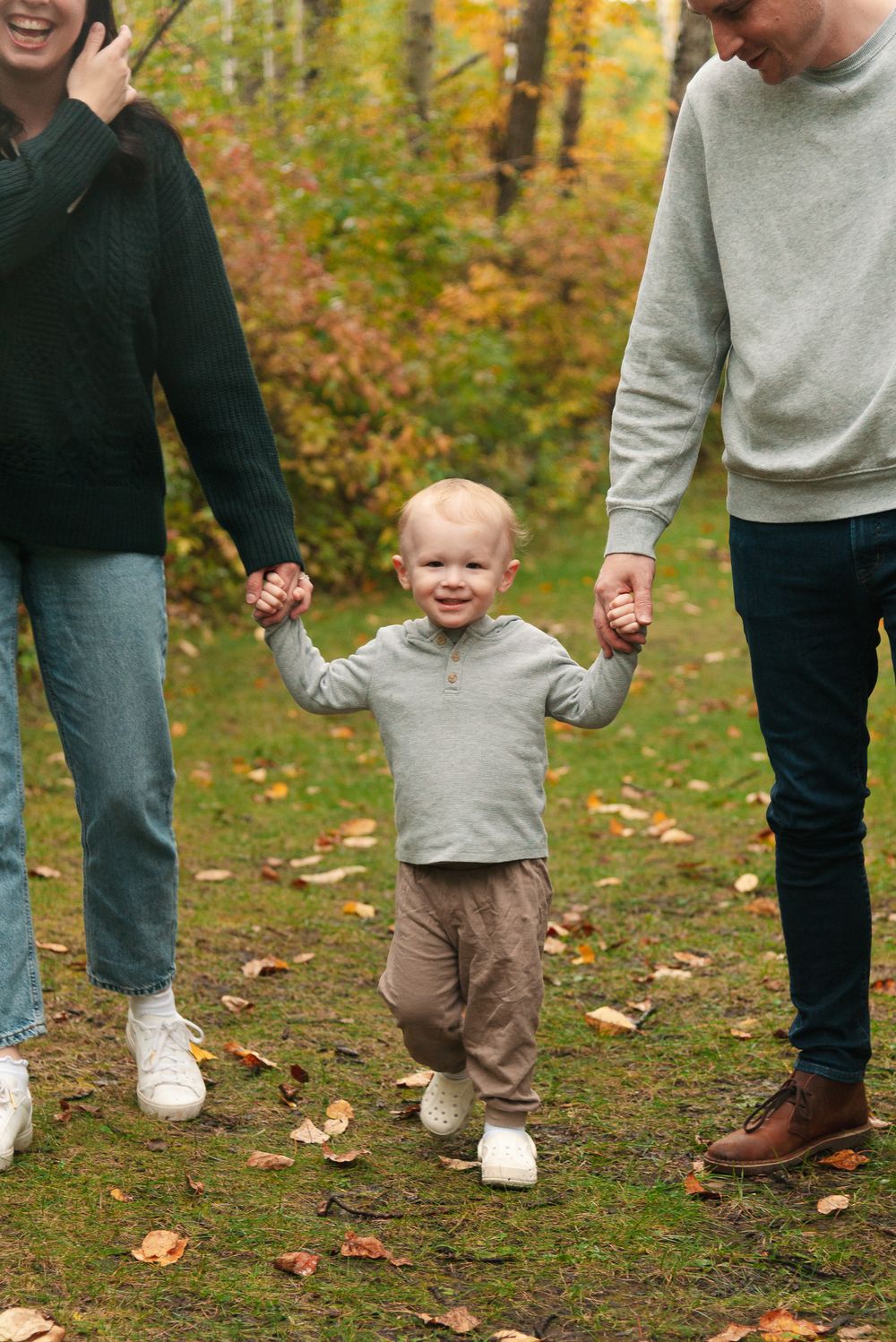 A toddler walks holding hands with parents on a leaf-covered forest path.